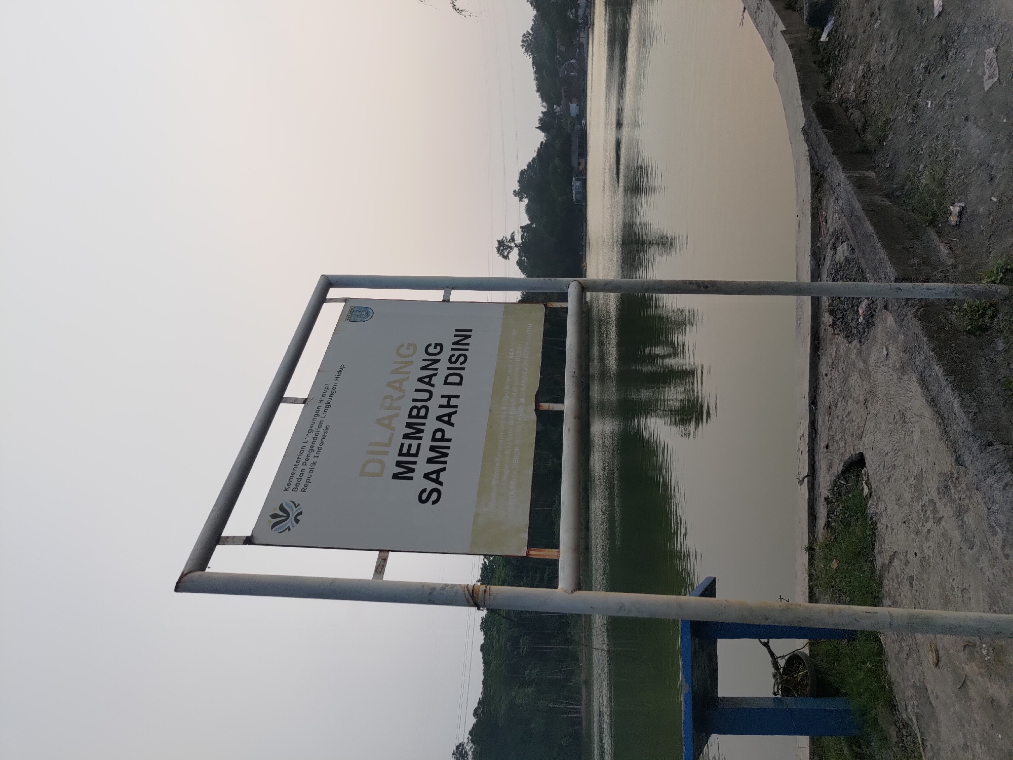 This is a medium shot, eye-level, outdoor photograph of a lake in Depok, Indonesia, taken from a concrete embankment. The sky is overcast and a pale yellow, suggesting either dawn or dusk. The lake occupies the majority of the frame, with its surface reflecting the muted light. On the left side of the image, a blue bench with a concrete base is visible, with a dark, circular object, possibly a planter or trash receptacle, resting on it. A white sign is mounted on a metal frame in the foreground, positioned slightly to the left of center. The sign has Indonesian text, with the prominent phrase "DILARANG MEMBUANG SAMPAH DISINI" meaning "DO NOT DUMP TRASH HERE". Above this, there is text indicating it is from the "Kementerian Lingkungan Hidup/ Badan Pengendalian Lingkungan Hidup Republik Indonesia" (Ministry of Environment/ Environmental Control Agency of the Republic of Indonesia). The background features a dense line of trees on the far bank of the lake, with some structures and a power line visible above them. The embankment in the foreground is rough and uneven, with patches of grass and scattered debris. The overall scene conveys a sense of quietness and a focus on environmental preservation.