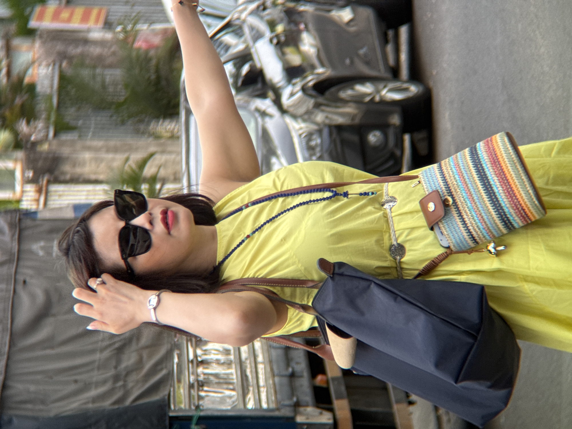 A woman in a yellow dress is standing on a street in Nha Trang, Vietnam, with a truck behind her and cars in the background. She has her hand up to her head, wearing sunglasses, and a necklace. She is carrying a large dark blue tote bag and a small colorful bag is attached to her belt. She is pointing with her right arm.