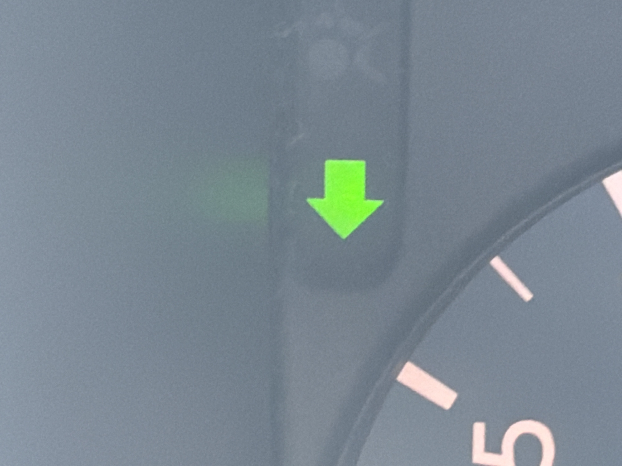 The image shows a green arrow pointing downwards illuminated on a vehicle dashboard. To the right of the arrow, a portion of a speedometer is visible, with the number "5" partially shown. The background appears to be a gray surface. There are no people in the frame. The location in the city of Izhevsk, Russia cannot be determined from this image.