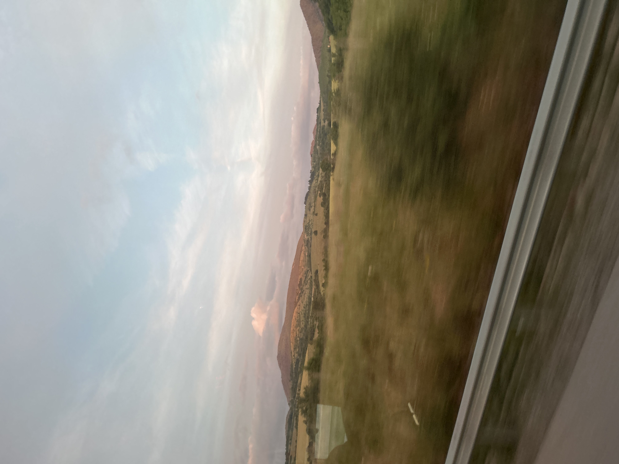 The image depicts a landscape view from a moving vehicle, likely a train or car, looking out a window. In the foreground, a blurred metal railing runs diagonally across the bottom of the frame. Beyond the railing, a field of green and brown grass is visible, also exhibiting motion blur. In the mid-ground, rolling hills covered in grass and scattered trees stretch across the scene. The hills are bathed in the soft, warm light of either sunrise or sunset, casting a golden hue on their slopes. Further in the background, more distant hills and mountains are visible, with a pinkish-orange tint. The sky above is a pale blue with wispy clouds tinged with pink and purple, characteristic of twilight. A solitary, small cloud with a distinct pink glow is present over the main hill in the mid-left. The overall impression is one of a rural landscape during a transitional time of day. The location is identified as Osikovitsa, Bulgaria.