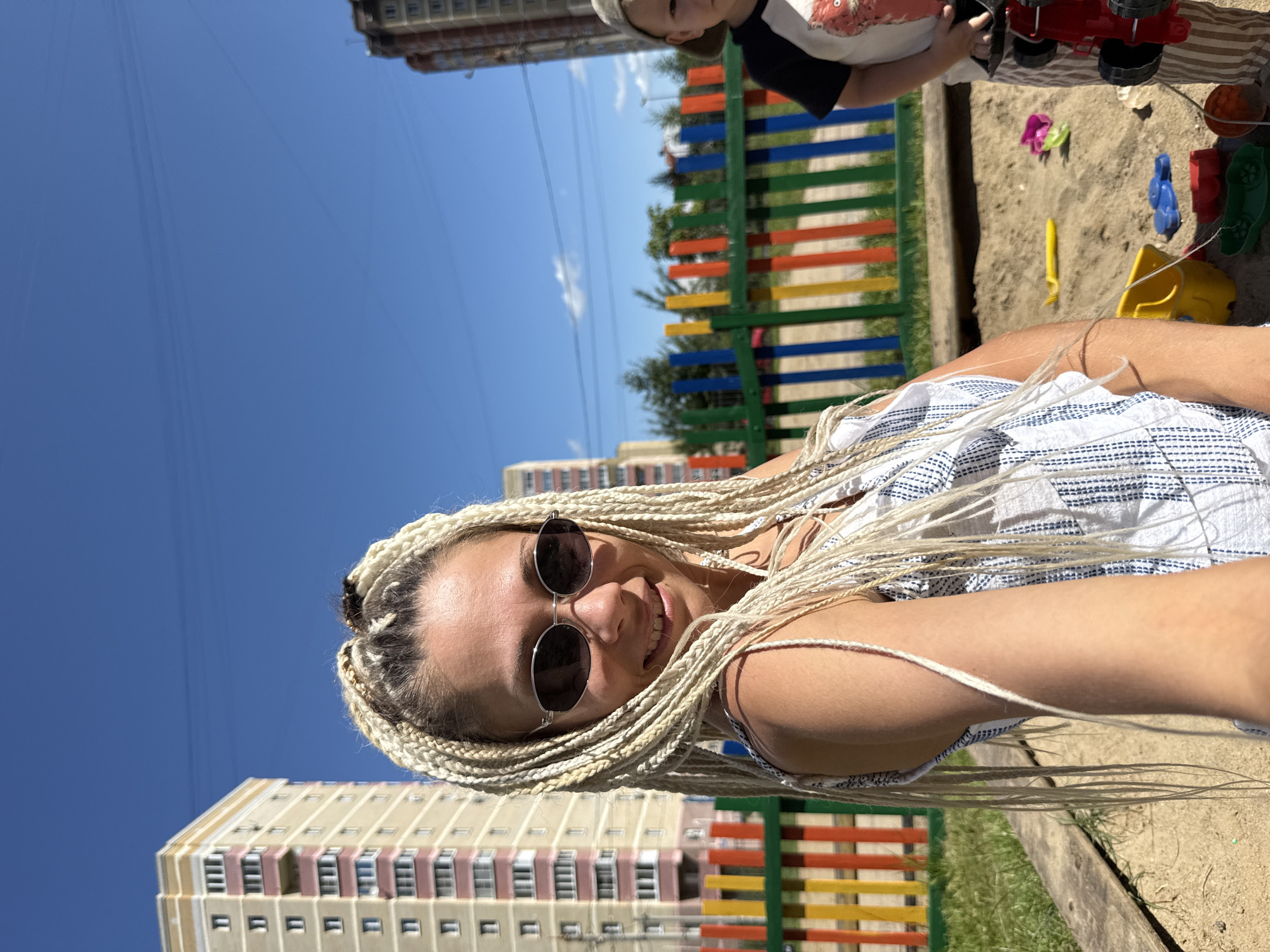 In Rusanovo, Russia, on a bright, sunny day, a woman with blonde braids is taking a selfie in a children's sandbox. She is wearing sunglasses and a white and blue striped outfit. Behind her, a colorful fence separates the sandbox from a grassy area and several apartment buildings. A young child, partially visible, stands to her right, holding a red toy. The sandbox contains various plastic toys and a yellow shovel.