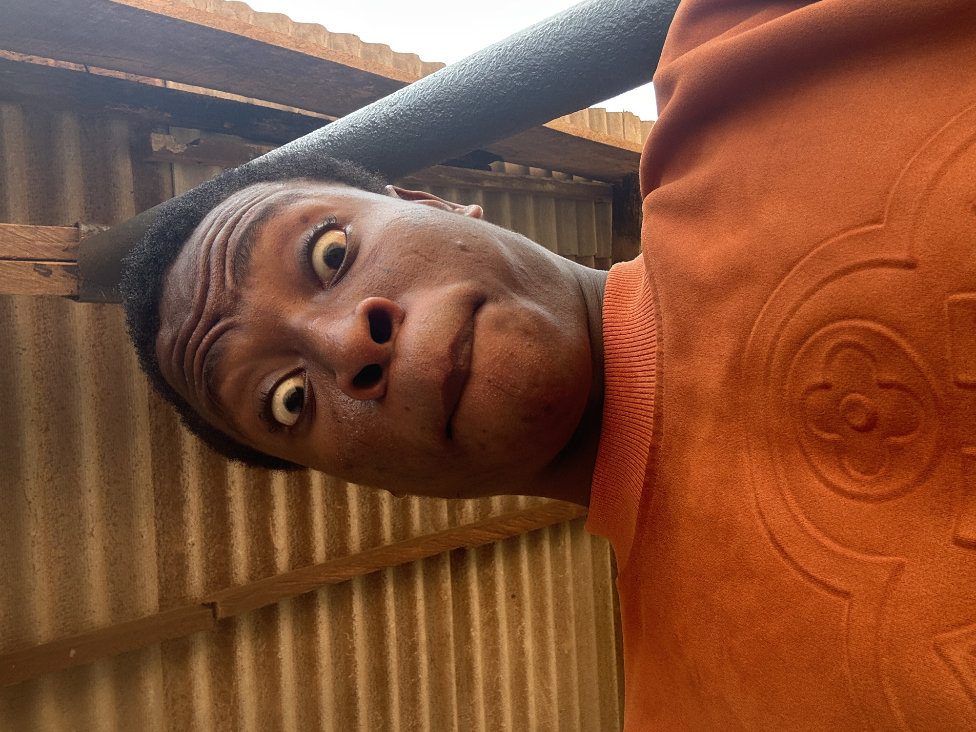 A low-angle photograph depicts a person with dark skin and short, dark hair. The person's eyes are wide and appear startled, and their facial expression is somewhat exaggerated with a furrowed brow and slightly opened mouth. The person is wearing a burnt orange sweatshirt with an embossed pattern on the right shoulder. The background consists of corrugated metal, wooden beams, and a black, cylindrical object overhead. The location appears to be an outdoor or semi-outdoor structure. The city and specific location are unknown.