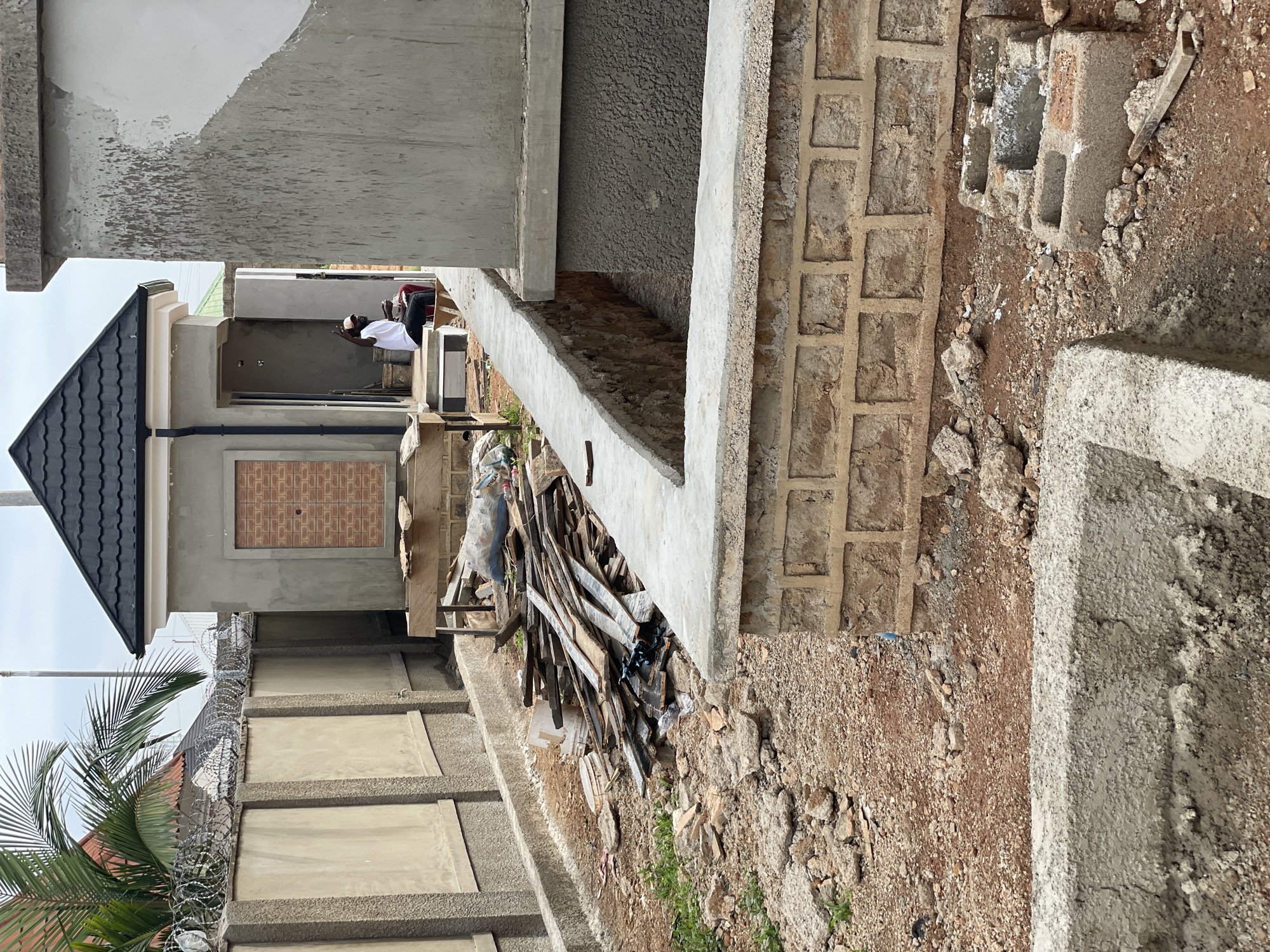 In Bar Gada, Nigeria, a construction site is depicted with several unfinished structures. A palm tree is visible to the left, with a concrete wall topped with barbed wire fencing. A small building with a dark, tiled roof and a brick-patterned door is in the background, with an open doorway revealing a person sitting inside. To the right, a larger, unfinished concrete structure is partially visible. The ground is covered in red dirt, gravel, and debris, including a pile of discarded wooden planks. Several low concrete walls and foundations are in various stages of construction. The sky is overcast.