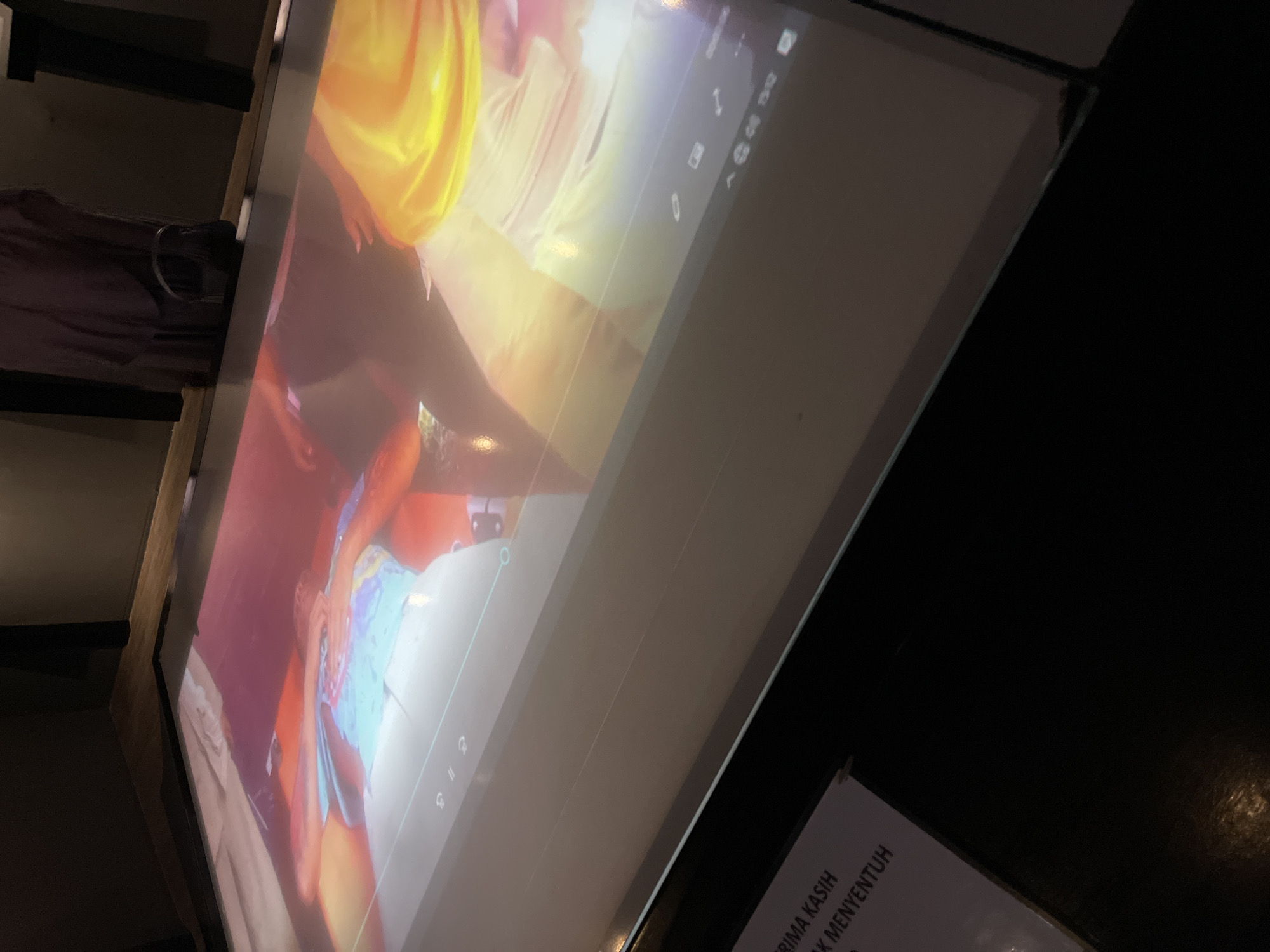 A projected image of people is displayed on a horizontal surface, likely a table, in a dimly lit room. The projected image shows at least two individuals, one in the foreground wearing a brightly colored shirt, and another person in the background. The projected image also displays user interface elements at the bottom, including playback controls for a video or presentation. To the left of the projected surface, a white sign with black text is partially visible, displaying the words "TERIMA KASIH" and "TENTUKAN". This suggests the location is in Banda Aceh, Indonesia, as "Terima Kasih" is Indonesian for "Thank You" and the context implies a visitor or service interaction. The environment appears to be indoors, possibly a museum, gallery, or public space, with dark walls and furniture.