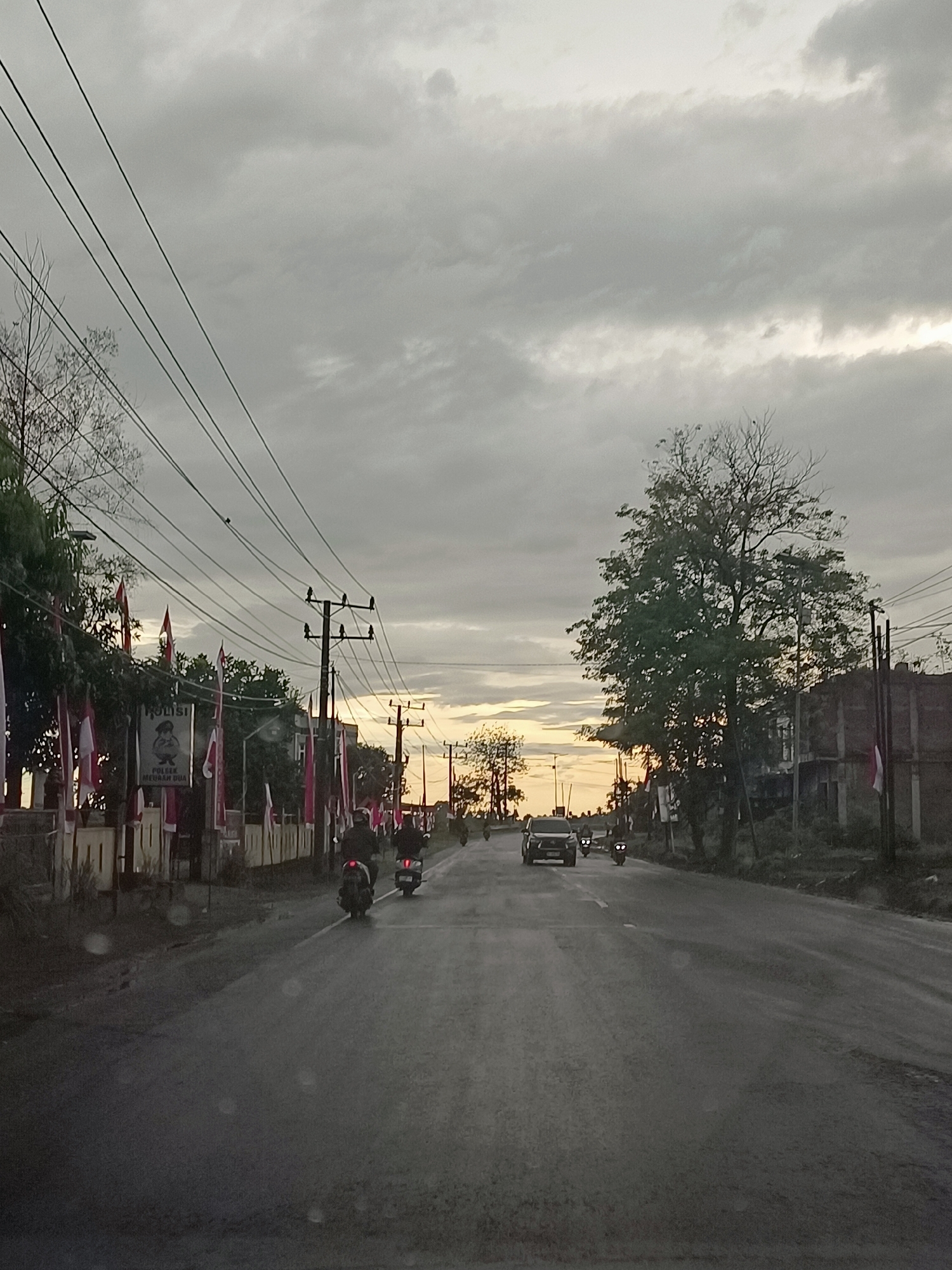 The image depicts a road scene in Meunasah Bie, Indonesia, during overcast conditions at dusk. A paved road runs through the center of the frame, with traffic present. Several motorcycles are seen on the road, with two in the foreground and more further down. A dark-colored car is also traveling in the same direction as the motorcycles. On the left side of the road, there is a building with a sign that reads "POLISI" (police) and a depiction of a police officer. This building is adorned with numerous red and white flags, possibly indicating a national celebration or event. Trees are visible on both sides of the road, some with sparse foliage, suggesting the presence of deciduous trees or the season. Utility poles with multiple power lines stretch across the upper portion of the image. The sky is filled with heavy, gray clouds, with a hint of lighter sky and potential sunset glow peeking through on the horizon in the distance. The overall atmosphere is muted, with the lighting suggesting the end of daylight.