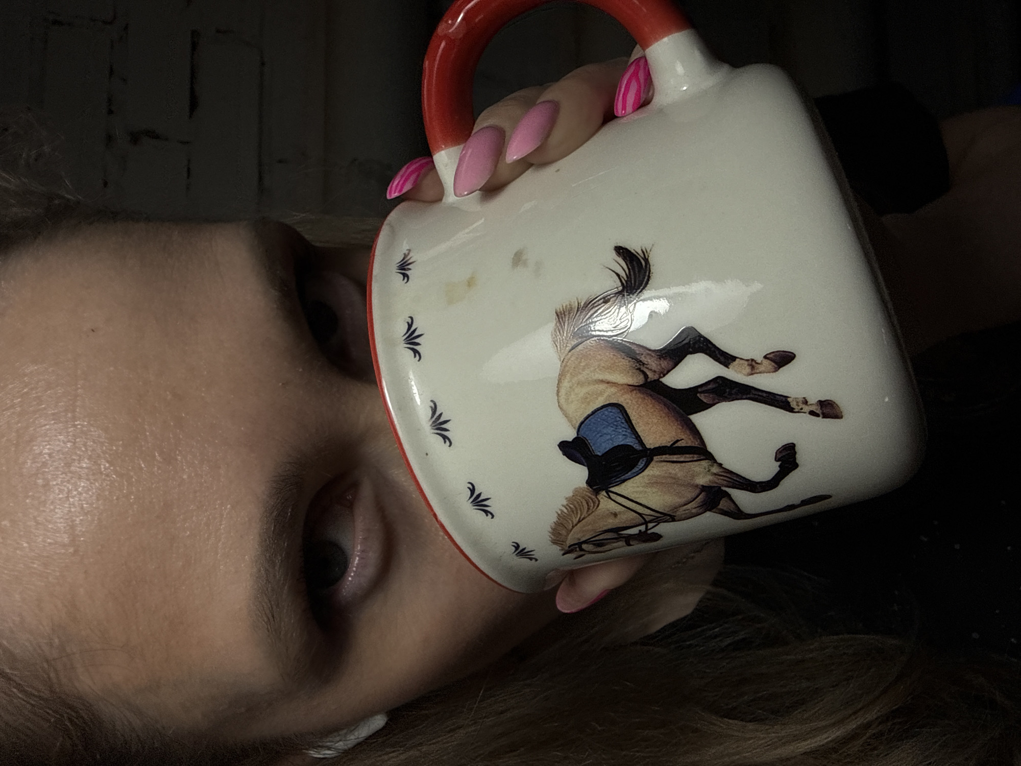 A person with light skin and light brown hair is holding a white mug with a red handle. The mug has a horse and saddle design on it. The person has pink manicured nails. The location is unspecified, and there is no indication that it is in the city of Chaikovsky, Russia. The background is dark and partially obscured.