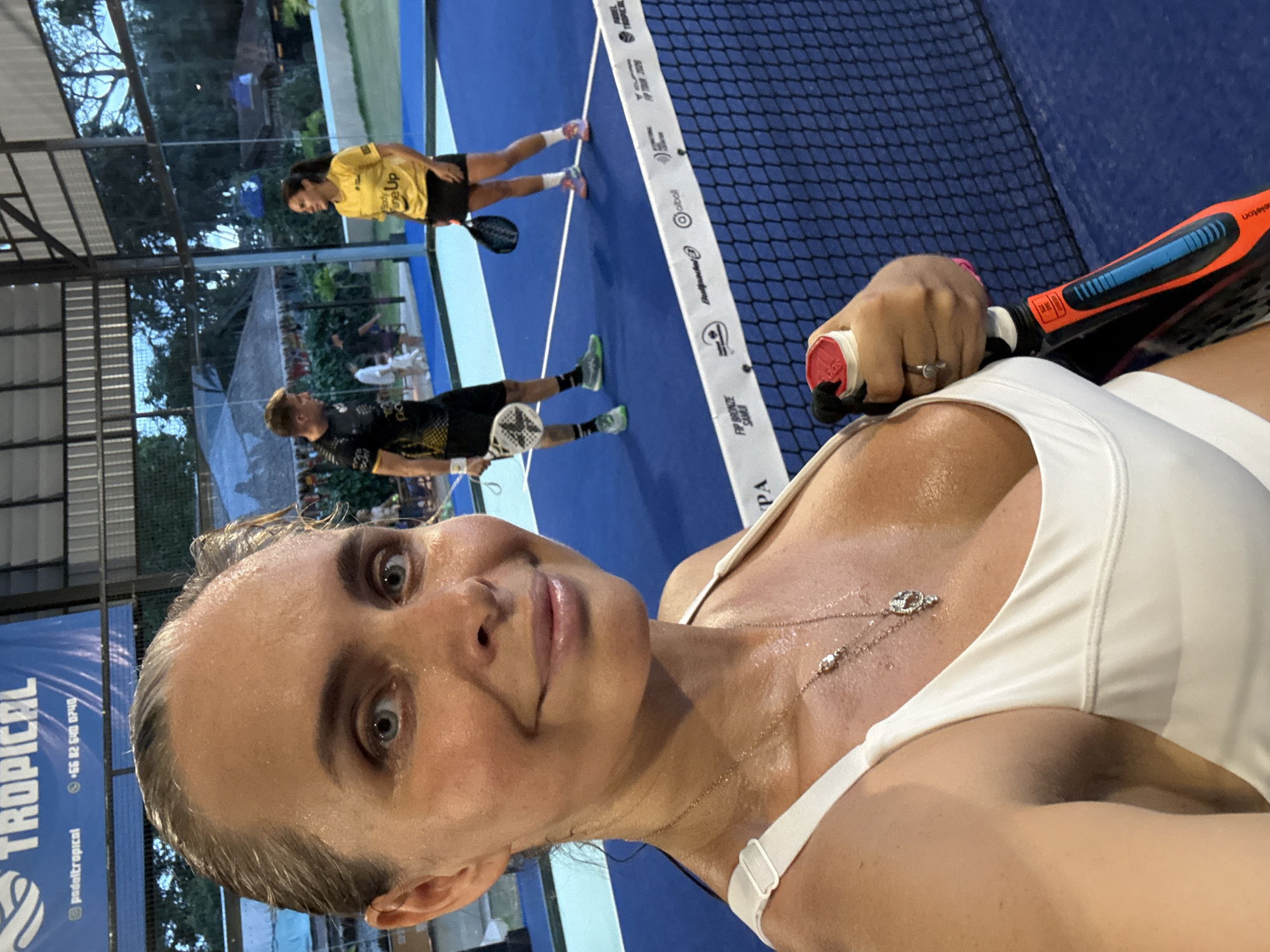 A close-up, front-facing selfie shows a woman with blonde hair pulled back, sweating profusely, wearing a white tank top and a necklace. Her makeup appears dewy, suggesting physical exertion. In the background, a man in a black and yellow shirt and a woman in a yellow shirt and black skirt are playing padel on a blue court. They are holding padel rackets. The scene is indoors, with a roof overhead and glass walls. The Padel Tropical logo is visible on a banner in the background, with the text "padeltropical.com" and a phone number. The location is specified as Ban Bang Rak, Thailand. The court is marked with white lines and a net separates the players.