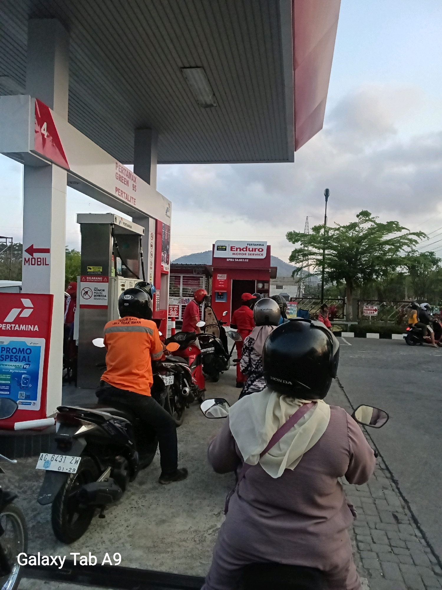 The image depicts a gas station in Trenggalek, Indonesia, likely during the late afternoon or early evening given the soft lighting. In the foreground, several people on motorcycles are queuing to refuel. One person wearing an orange shirt and black helmet is prominent, seated on a black motorcycle with license plate AG 6431 ZW. Another individual in a lilac-colored outfit and white hijab is also on a motorcycle, facing away from the camera. A red Pertamina sign with the company logo is visible on the left, advertising "PROMO SPESIAL" for Pertamax 95.
Behind the first motorcycle, other individuals and motorcycles are lined up at a Pertamina fuel pump. A red structure with the Pertamina Enduro Motor Service logo is situated further back. Gas pumps are clearly visible, with attendants in red uniforms attending to customers. The environment shows a paved ground with some cobblestone detailing on the edges, and a canopy structure covering the fuel pumps. In the background, trees and hills are visible under a cloudy sky. The overall scene captures a routine moment at a local gas station in Trenggalek.