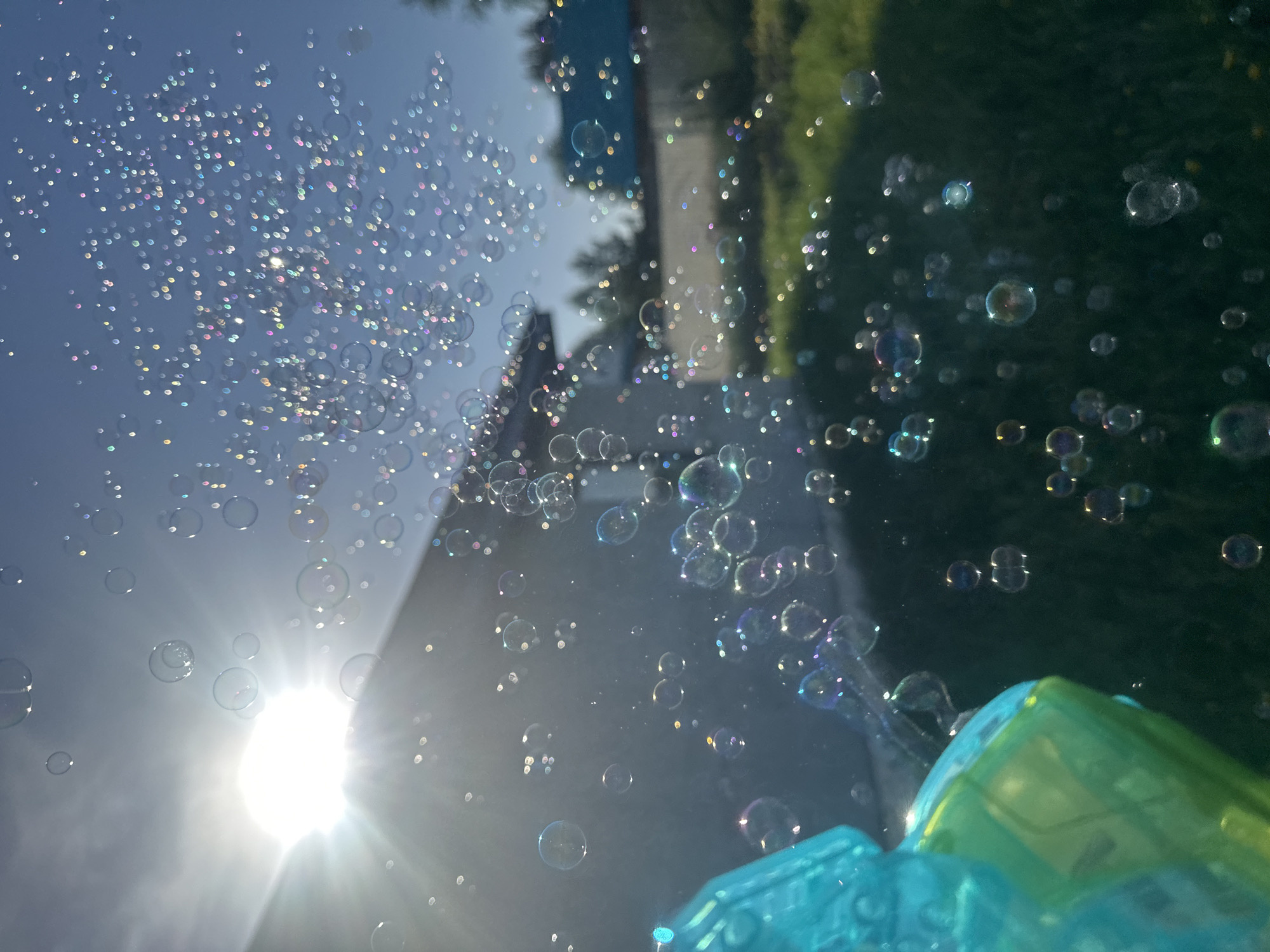 The image shows numerous soap bubbles in the air against a backdrop of a sunny sky and partial view of green vegetation and a building. In the foreground, portions of a blue and green plastic toy, possibly a bubble gun, are visible. The sun is shining brightly from the lower left corner, creating a lens flare effect and illuminating the iridescent bubbles. The location is not discernible as Kharnavy, Russia, from the visual data provided.