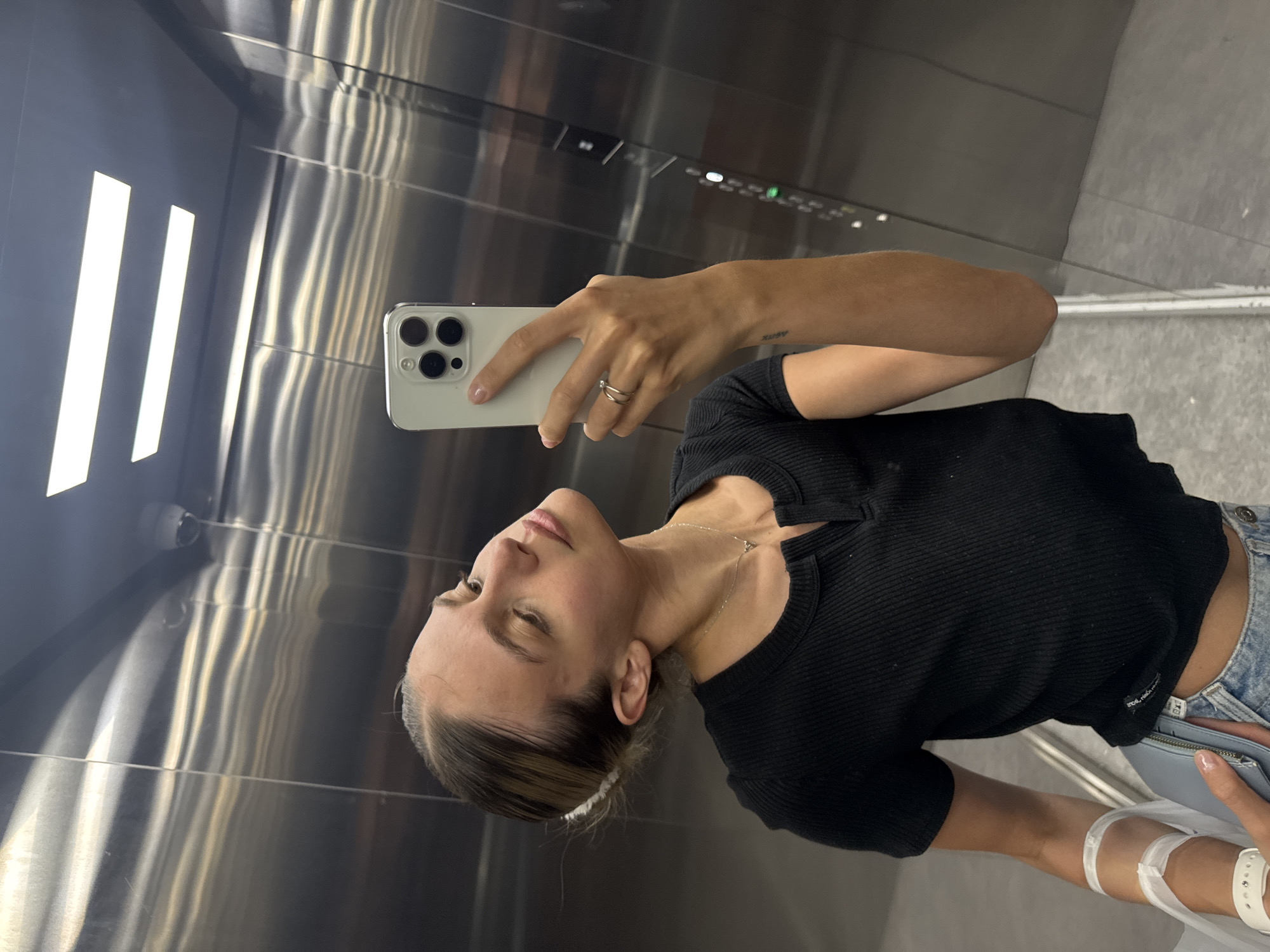 A woman is taking a selfie in an elevator. She is holding a white smartphone with multiple camera lenses. The woman is wearing a black shirt and blue jeans, and has a medical wristband on. The interior of the elevator has stainless steel walls and ceiling, with integrated lighting panels. The location is in the city of Unknown, Unknown.