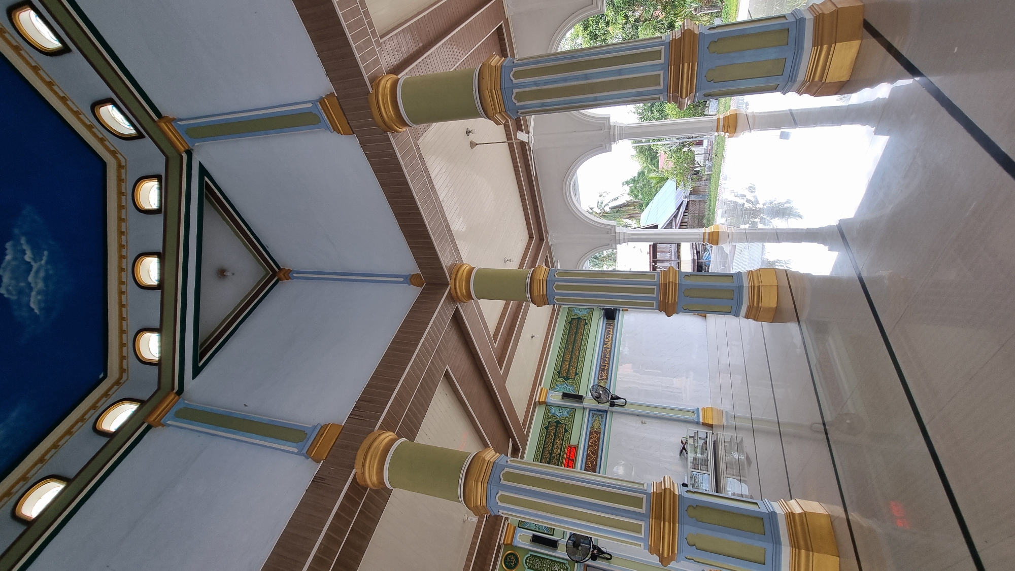 This is a low-angle, wide shot of the interior of a mosque, taken from a tiled floor reflecting the scene. Four large, decorative pillars, painted in pastel blue and green with gold accents at the top and bottom, dominate the foreground. The pillars support a ceiling with a brown, grid-like pattern. Above this, the ceiling transitions to a light blue with painted white clouds, and arched windows with orange-tinted glass are visible along the upper edge of the walls. The walls themselves are light blue and white. In the background, through an open archway, glimpses of lush green trees and some buildings can be seen, suggesting an outdoor environment. The polished floor is a light beige with two thin black lines running parallel to each other, creating a sense of perspective. There are no people visible in the image. The overall impression is one of serene and ornate architectural design. The location is identified as Meunasahlhok, Indonesia.
