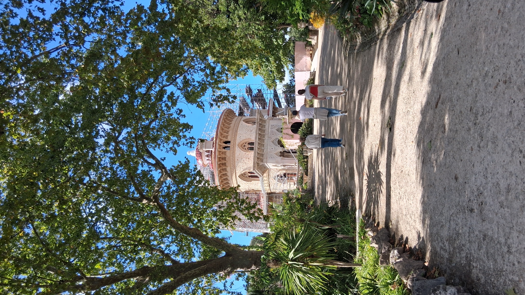 In Sintra, Portugal, within the parish of Santa Maria e São Miguel, São Martinho e São Pedro de Penaferrim, three individuals walk along a gravel path towards a distinctive, circular building adorned with scaffolding. The building features ornate stonework, arched windows, and a red-tiled dome. Lush green trees with dense foliage frame the scene, casting dappled shadows on the path. The sky is clear and blue. The path is bordered by rocks and greenery on the left side. The people, viewed from behind, are casually dressed.