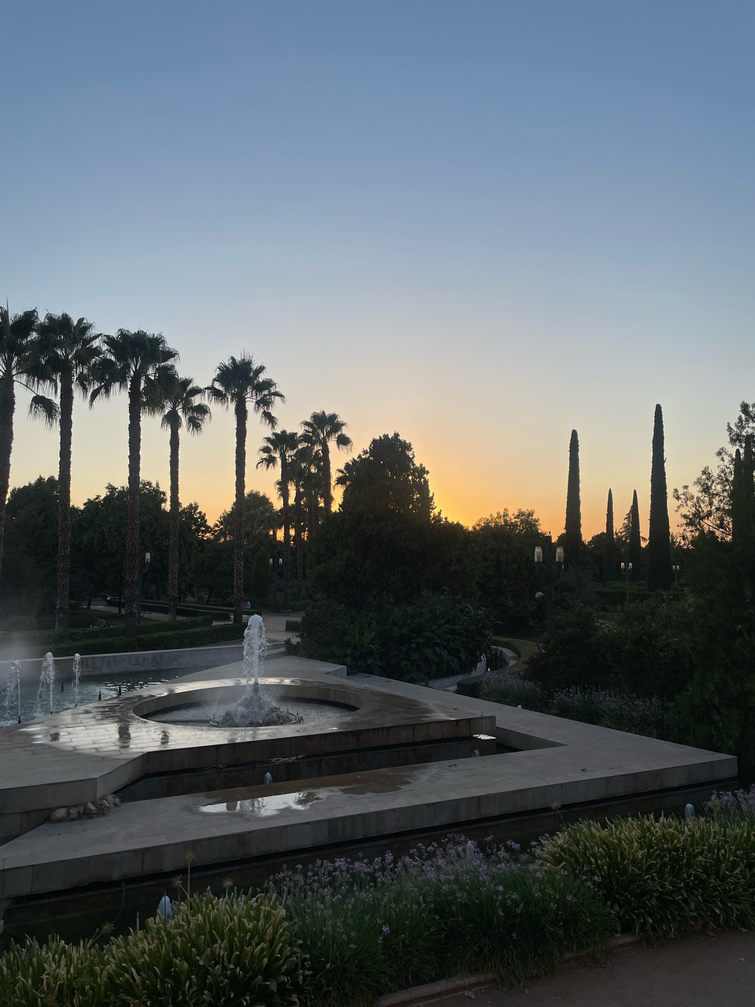This image depicts a sunset scene in the Carmen de los Mártires gardens in Granada, Spain. The foreground features a tiered fountain with water jets and reflections. Palm trees are prominent on the left side of the image, with a line of cypress trees visible on the right. The sky is a gradient of blue, transitioning to a soft orange and yellow where the sun is setting behind trees and other foliage. Several streetlights are visible amongst the greenery. The ground in the foreground appears to be a path or dirt area. The overall atmosphere is serene, with the warm hues of the sunset contrasting with the cooler tones of the sky and the shadows cast by the trees.
