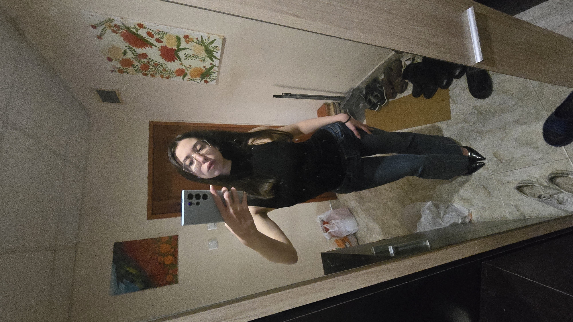 A mirror selfie shows a woman with long, dark hair, wearing glasses, a black top, and jeans. She's holding a phone to take the photo. The mirror reflects a hallway with a door, artwork on the wall, and a collection of shoes on the floor. The background is a residential interior, with tiled flooring. While the location in Stara Zagora, Bulgaria is unconfirmed in the image, the setting is most likely a private residence.