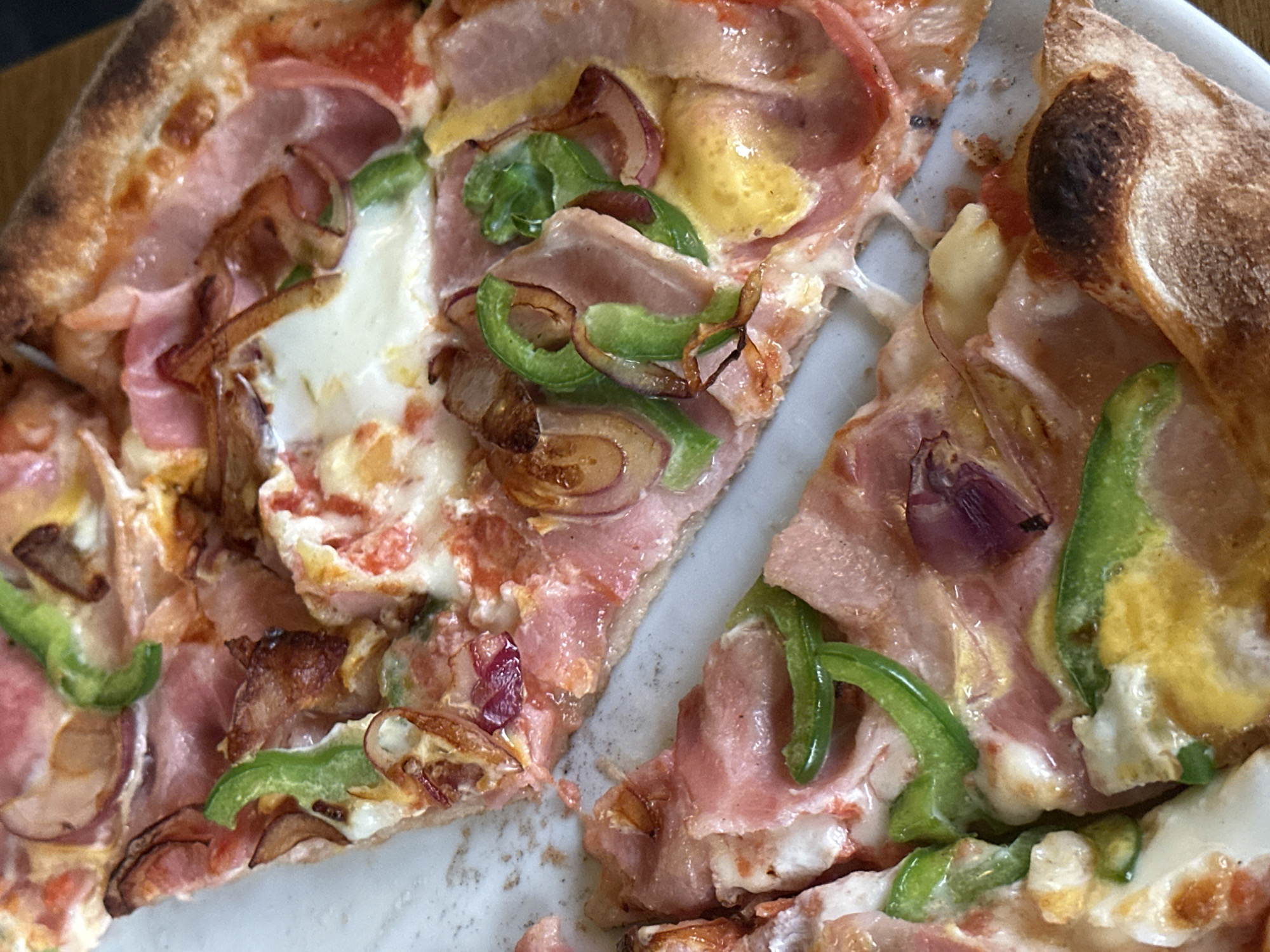 The image shows a close-up of a pizza with toppings including ham, green peppers, and caramelized red onions. The pizza is cut into slices. The crust is golden brown, with some darker burnt spots. The setting or location in Sofia, Bulgaria, is not discernible from the provided image.