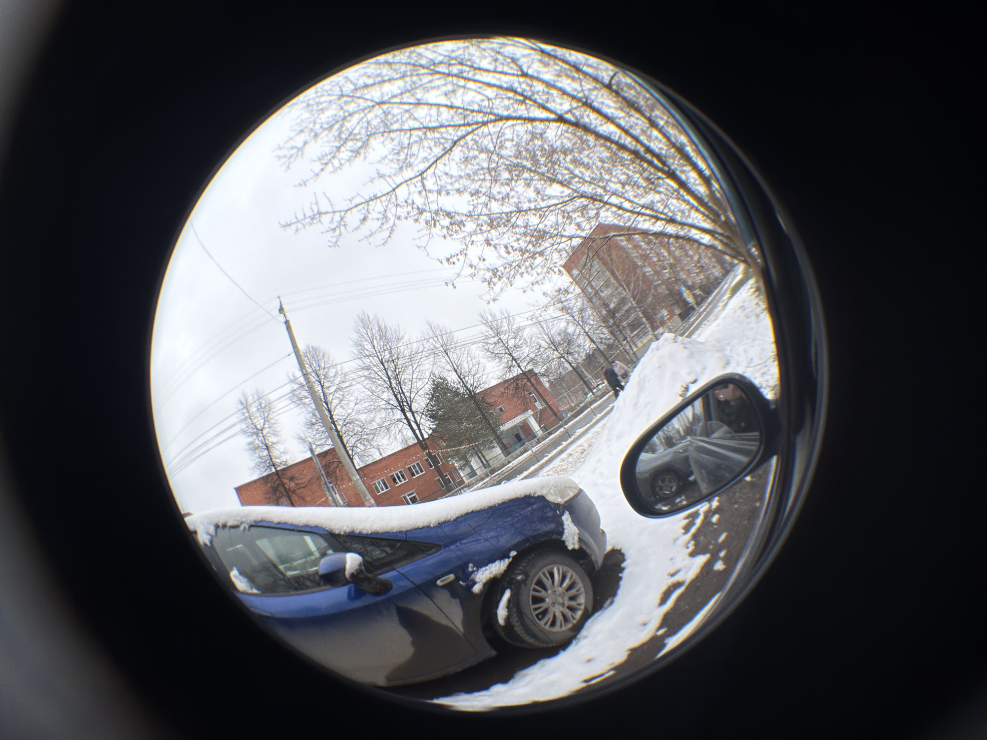 The image, taken with a fisheye lens, shows a winter scene in Izhevsk, Russia. A blue car is parked in the snow, with snow accumulated on its roof. In the car's side mirror, a person's reflection is visible. A brick building is in the background, partially covered in snow. Bare trees and power lines are visible in the sky.