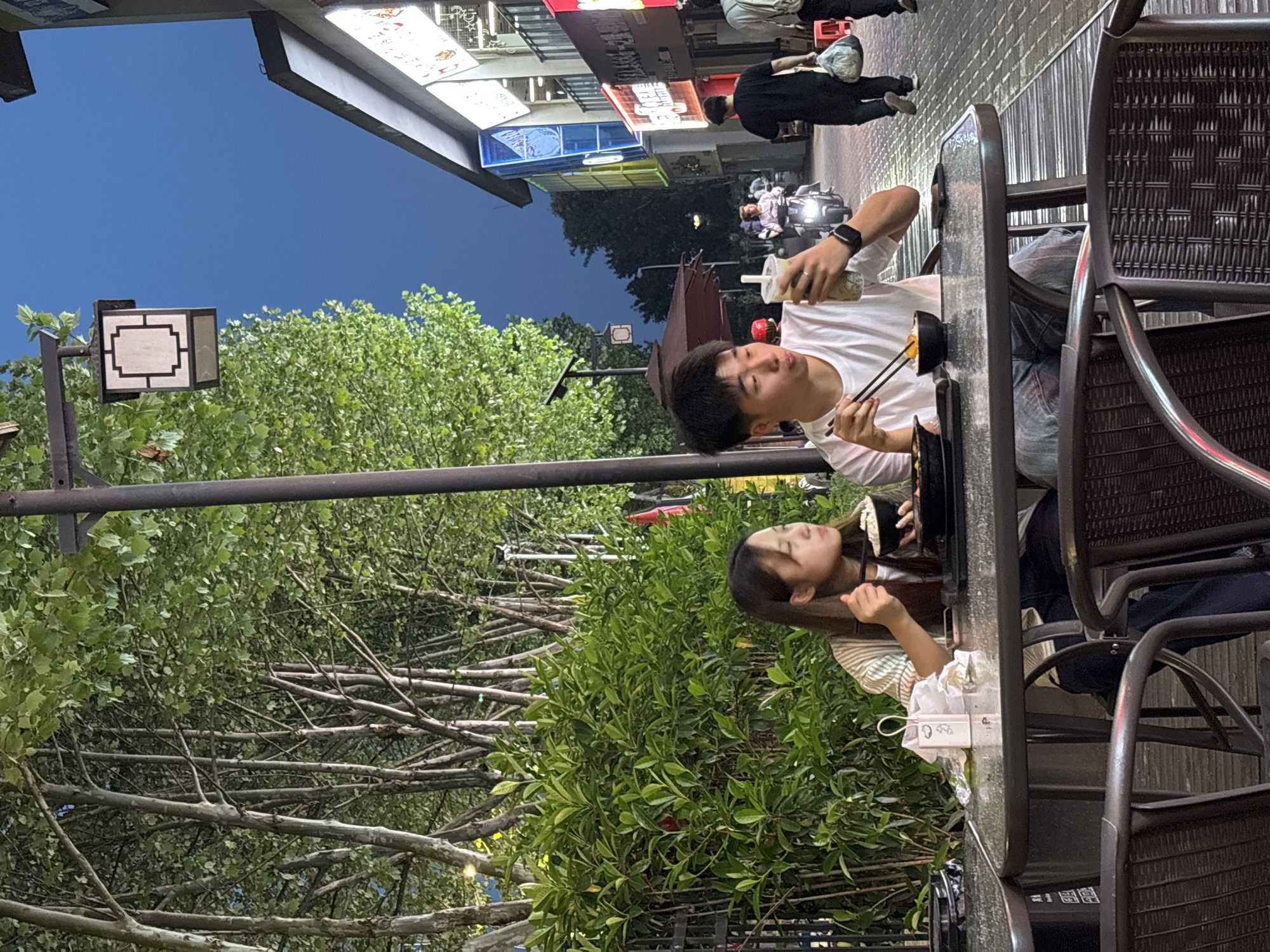Here is a description of the image:
The image is taken at night in what appears to be Jiangjin, China. A young man and a young woman are seated at an outdoor table at a restaurant. The man is holding chopsticks in his right hand, and a beverage with a straw in his left. The woman is leaning on the table and looking in the direction of the man. There is foliage of some trees in the background on the left, and some building lights on the right. A few pedestrians, and a motorbike, can also be seen in the background on the right.