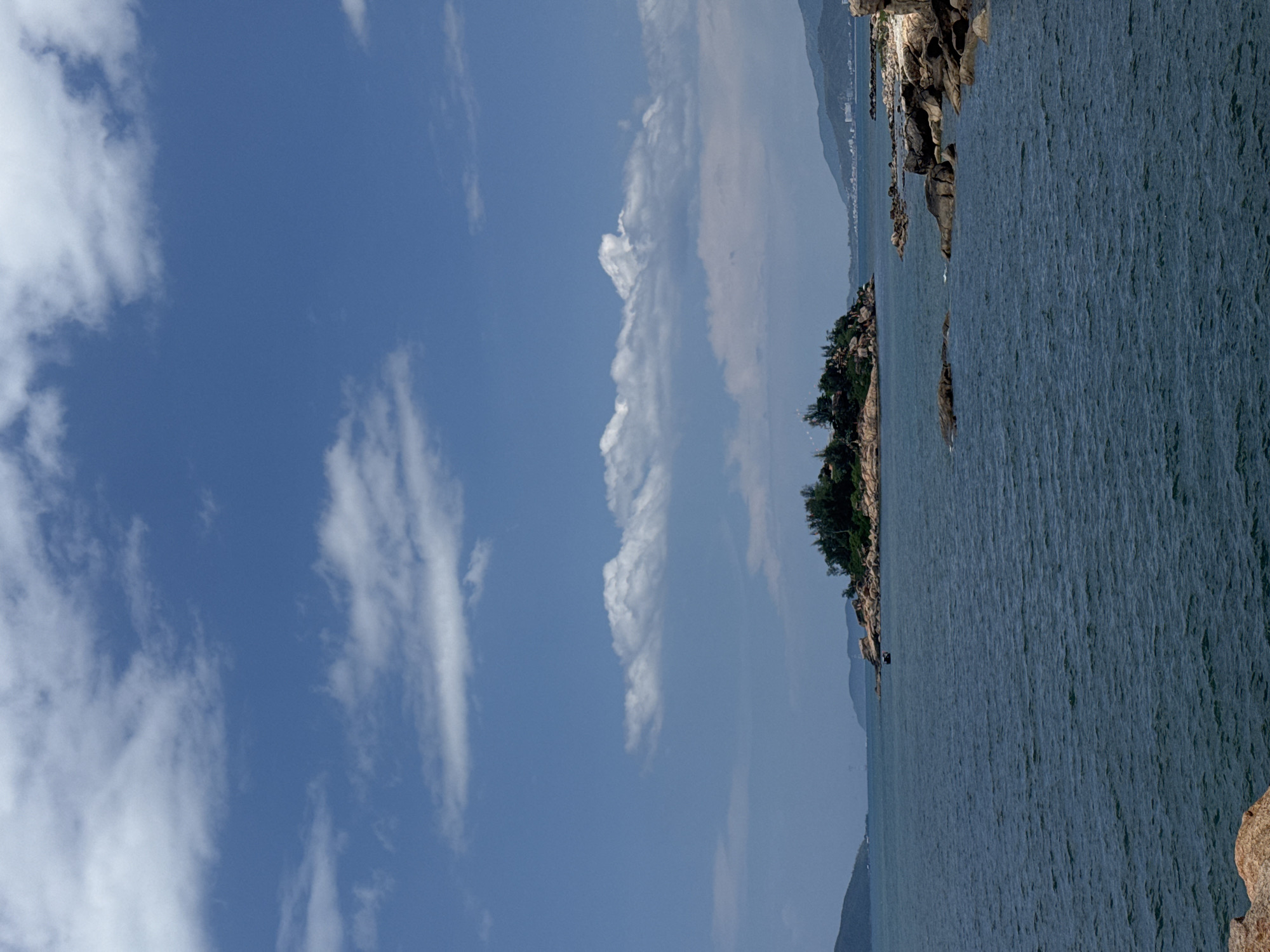 This landscape photograph captures a serene coastal scene under a bright, expansive blue sky. The upper portion of the image is filled with a clear, deep blue sky, adorned with scattered white clouds ranging from fluffy cumulus to wispy cirrus formations. The lighting indicates daytime, likely late morning or early afternoon, with clear and sunny weather conditions.
The mid-ground reveals a rugged coastline, featuring rocky outcrops and small islands that extend into the water. These land formations are partially covered with vibrant green vegetation, including trees and bushes, providing a lush contrast against the blues of the sky and sea. In the far distance, faint, hazy mountains or hills are visible, blending softly into the horizon.
The lower and right-hand side of the image (when oriented as a landscape) is dominated by a vast expanse of dark blue-green water, suggestive of the sea or ocean. The water's surface exhibits subtle texture from numerous small waves or ripples, implying a gentle current or breeze.
There are no people, animals, or prominent man-made structures visible in the scene, emphasizing the untouched natural beauty of the environment. The overall impression is one of tranquility and natural grandeur, typical of a beautiful coastal area. No visible text is present. This natural setting aligns with the provided location context of Bac Nha Trang Ward, Vietnam.