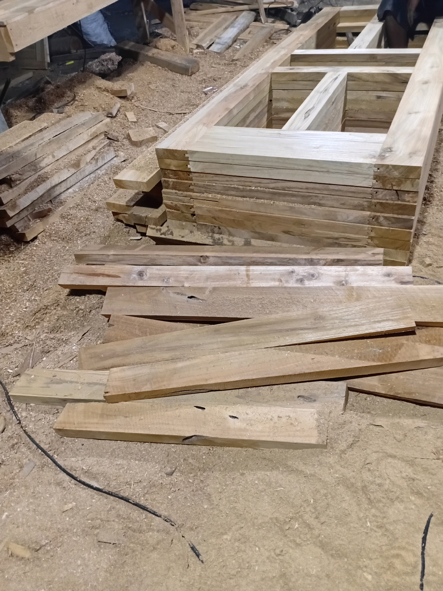 This image shows a woodworking workshop located in Playen, Indonesia. The workshop is filled with various wooden materials, including a stack of lumber and a partially assembled wooden frame, possibly for a door or window. Sawdust is scattered across the ground, indicating recent activity. The overall scene suggests a functional, working environment within a building, possibly a house or a dedicated workshop.