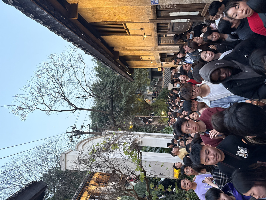 The image depicts a densely packed pedestrian street or alleyway within Nan'an District, China, at dusk or early evening. Hundreds of individuals, predominantly appearing to be of East Asian descent, fill the entire width of the thoroughfare, moving generally towards the background. Most are dressed in casual attire, with some wearing face masks.
On the right side of the street, a two-story building with a textured, yellow-ochre facade and a traditional dark tiled roof is visible. It features several rectangular windows with vertical wooden grates and multiple illuminated wall-mounted lamps. A dark wooden board with illegible writing is affixed to the wall, along with a white vertical sign bearing the number "117."
On the left side, a lighter-colored, possibly white or off-white, traditional-style building with a distinctive arched gateway is present. This building also has a dark tiled roof. Further into the background on the left, additional traditional structures are visible amidst lush green foliage.
Above the buildings and extending across the sky, a network of overhead utility wires is present, supported by a utility pole positioned behind the white arched structure. A large, bare deciduous tree with slender branches and some small red buds stands prominently against the light blue-grey twilight sky towards the right. Other trees with dense green foliage are visible on the hillsides in the background. Artificial warm yellow light emanates from the building lamps and some interior lights, contrasting with the fading natural light.