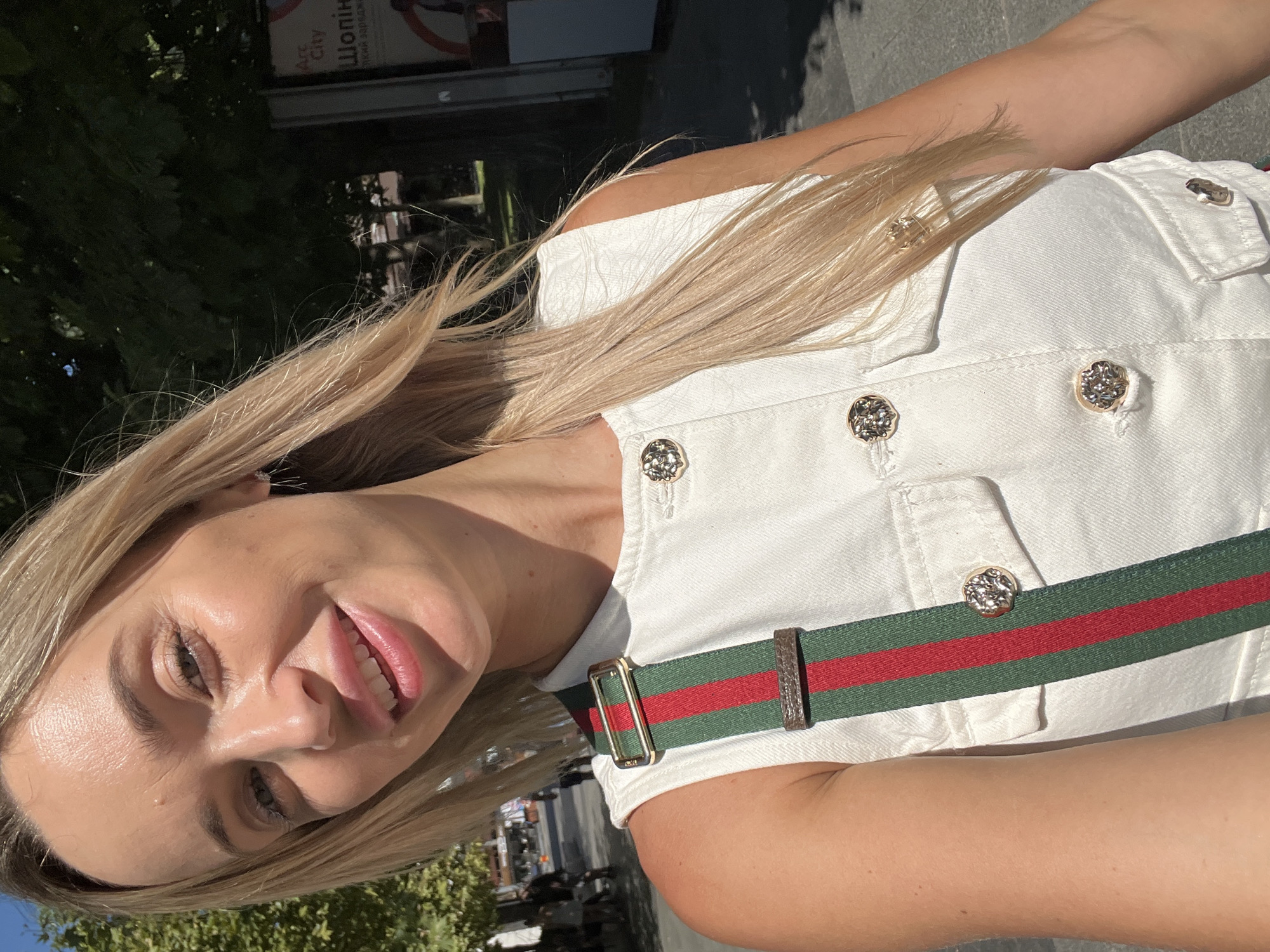A fair-skinned blonde woman, wearing a white sleeveless denim top with gold buttons and a green and red striped strap, takes a selfie outdoors in Odesa, Ukraine. Her blonde hair is windswept. The background shows trees, a sidewalk, and a billboard in Ukrainian. Sunlight casts shadows on the ground, indicating a daytime setting.