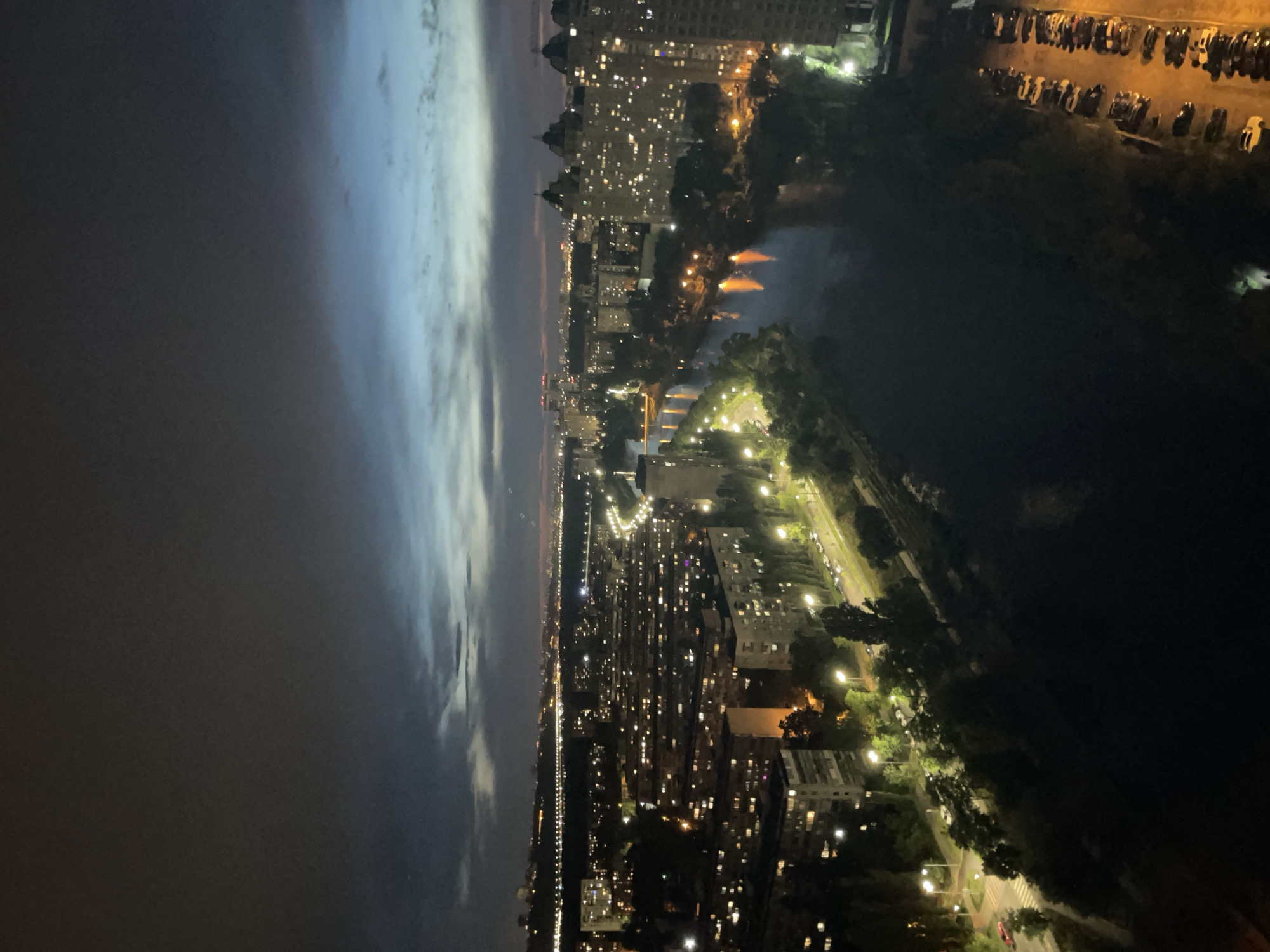 This is a nighttime aerial view of the Dnipro River in Kyiv, Ukraine, with the cityscape illuminated by artificial lights. The sky displays a gradient of dark grey at the top, transitioning to blues and hints of pink near the horizon, suggesting twilight or early dawn.
On the left side of the image, a dense urban landscape of apartment buildings is visible, their windows lit, creating a warm glow. Streetlights line the roads that wind through the residential areas. Trees are scattered throughout the city, their dark forms contrasting with the lights.
The Dnipro River flows through the center of the image, its dark waters reflecting the lights from the city. On the right bank of the river, more tall apartment buildings are present, also illuminated. Orange reflections of lights are visible on the river's surface in some areas. Along the embankment, trees and vegetation create a green buffer zone.
Further in the distance, a broader cityscape stretches across the horizon, with lights indicating more development and infrastructure, possibly including bridges or elevated roadways. On the far right of the foreground, a parking lot filled with cars is visible. The overall impression is of a large, bustling city at night, with the river acting as a central artery.
