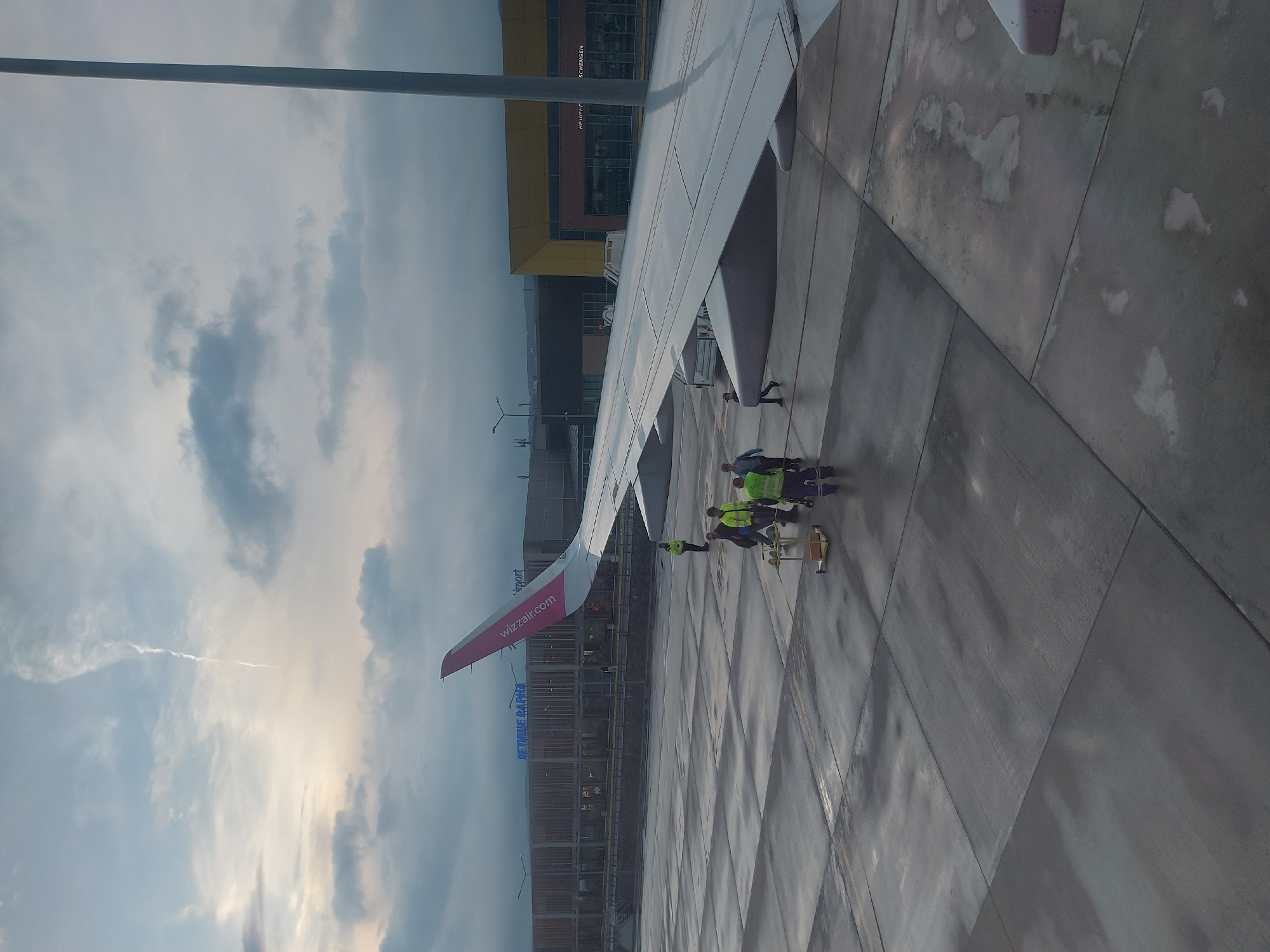 The image shows an airplane wing, presumably from a Wizzair plane, at an airport. The tail fin with the Wizzair logo is visible. A group of airport workers in bright yellow vests are working around the plane. The airport building is in the background, with signage in both English and Cyrillic letters, including the word "Airport" and "Аеропорт". The Cyrillic text includes "Летище Варна", suggesting the location is Varna Airport in Bulgaria, which is located near Aksakovo. The sky is cloudy. The ground appears to be a concrete surface of the apron.