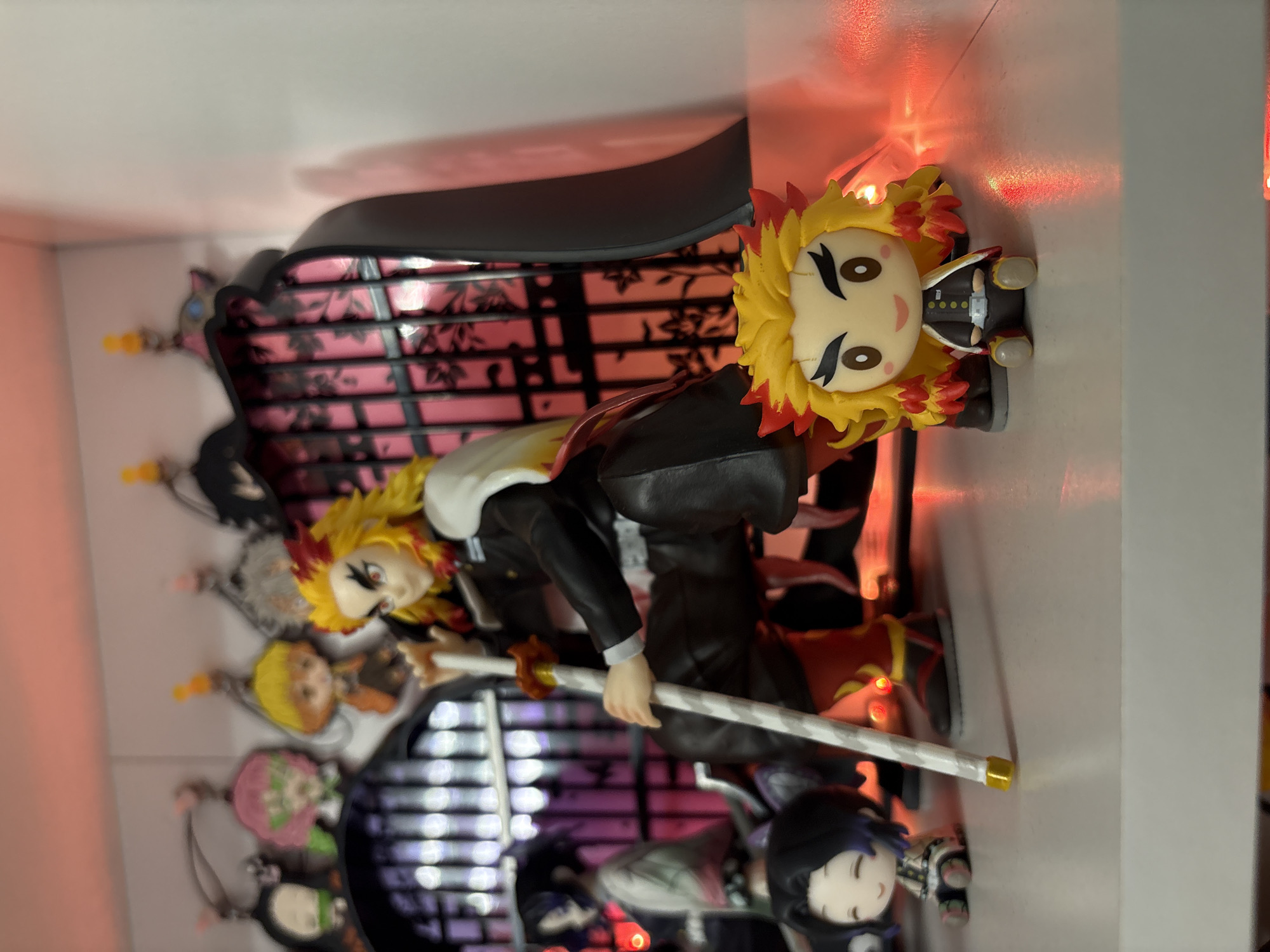 The image depicts an arrangement of anime figurines, specifically from the series "Demon Slayer." The central figure is Rengoku Kyoujurou, poised with his sword, in what appears to be a display case with a backdrop resembling a Japanese-style lattice with floral designs. Smaller chibi-style figures of other characters, including what appear to be Nezuko and Shinobu, are also present within the case. The backdrop is illuminated with purple and red lights.
The image doesn't provide information about the city of Plovdiv, Bulgaria.