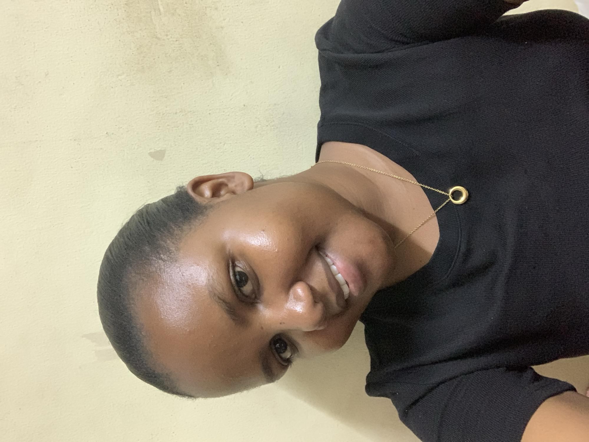 A dark-skinned woman smiles at the camera, wearing a black long-sleeved shirt and a gold necklace with a circular pendant. The background is a light yellow wall. The picture's location or a description of the city of Dar es-Salaam, Tanzania, cannot be determined from the image.