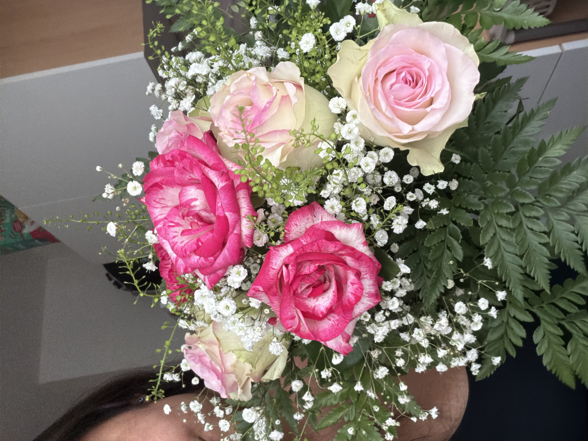 A close-up, vertical, medium shot shows a bouquet of roses and baby's breath flowers being held by a person whose face is partially visible on the left side of the frame. The person has dark hair and is wearing a dark top. The bouquet is composed of several roses in shades of pink and cream, with delicate white baby's breath filling the spaces between the larger blooms. Green ferns provide a leafy backdrop. The background is out of focus and appears to be an indoor setting with a neutral-colored wall and a hint of wooden paneling. No specific location in Duisburg, Germany can be identified from the visual information provided.