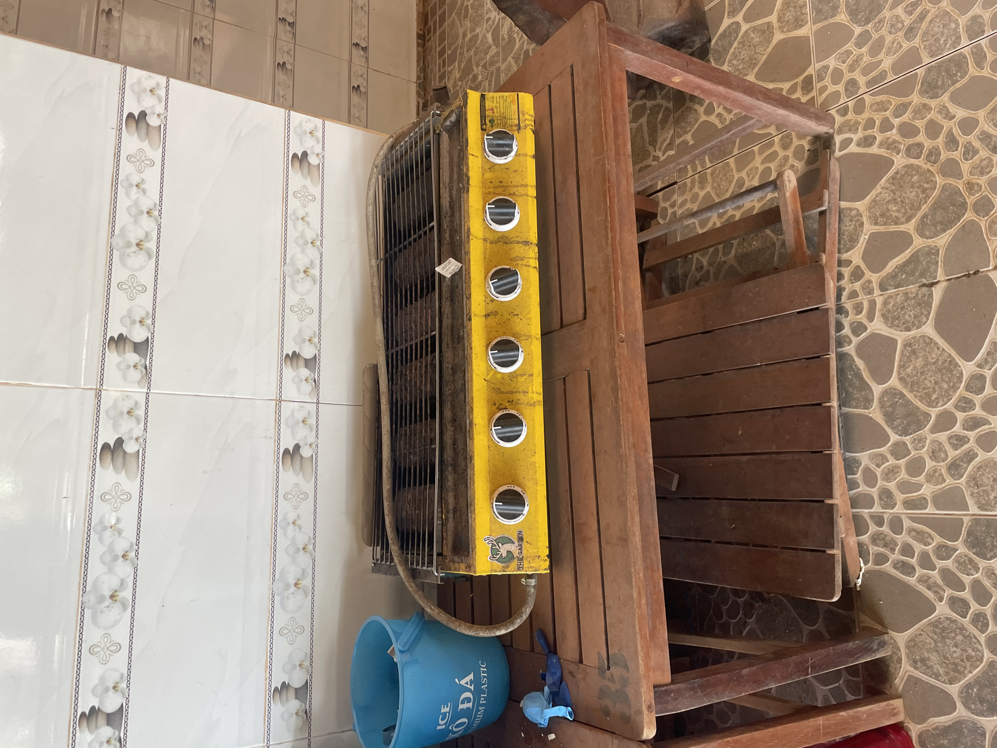 The scene depicts an outdoor or utility area in Chư Sê, Vietnam. A rectangular wooden slatted table, exhibiting signs of wear and featuring a partially detached slat on its lower shelf, occupies the central foreground. Resting on the table is a heavily used, grime-covered yellow gas grill with a black metal grate. The grill's front panel displays five black control knobs, a logo with a deer and the text "THE GARDEN," and a warning label on the right side. A tan gas hose connects to the left side of the grill. To the left of the table, a light blue plastic bucket rests on the tiled floor, bearing partially visible text "ICE ĐÁ PREMIUM PLASTIC." Beneath the bucket and near the table leg, several deflated blue latex objects, resembling balloons or gloves, are scattered on the floor. The background consists of a white tiled wall adorned with two horizontal decorative borders, each featuring white orchids, a four-leaf clover motif, and stacked stones. To the right, a section of a beige tiled wall with similar decorative bands is visible. The floor is covered with a pattern of interconnected, irregularly shaped light brown and gray tiles. No individuals are present in the image.