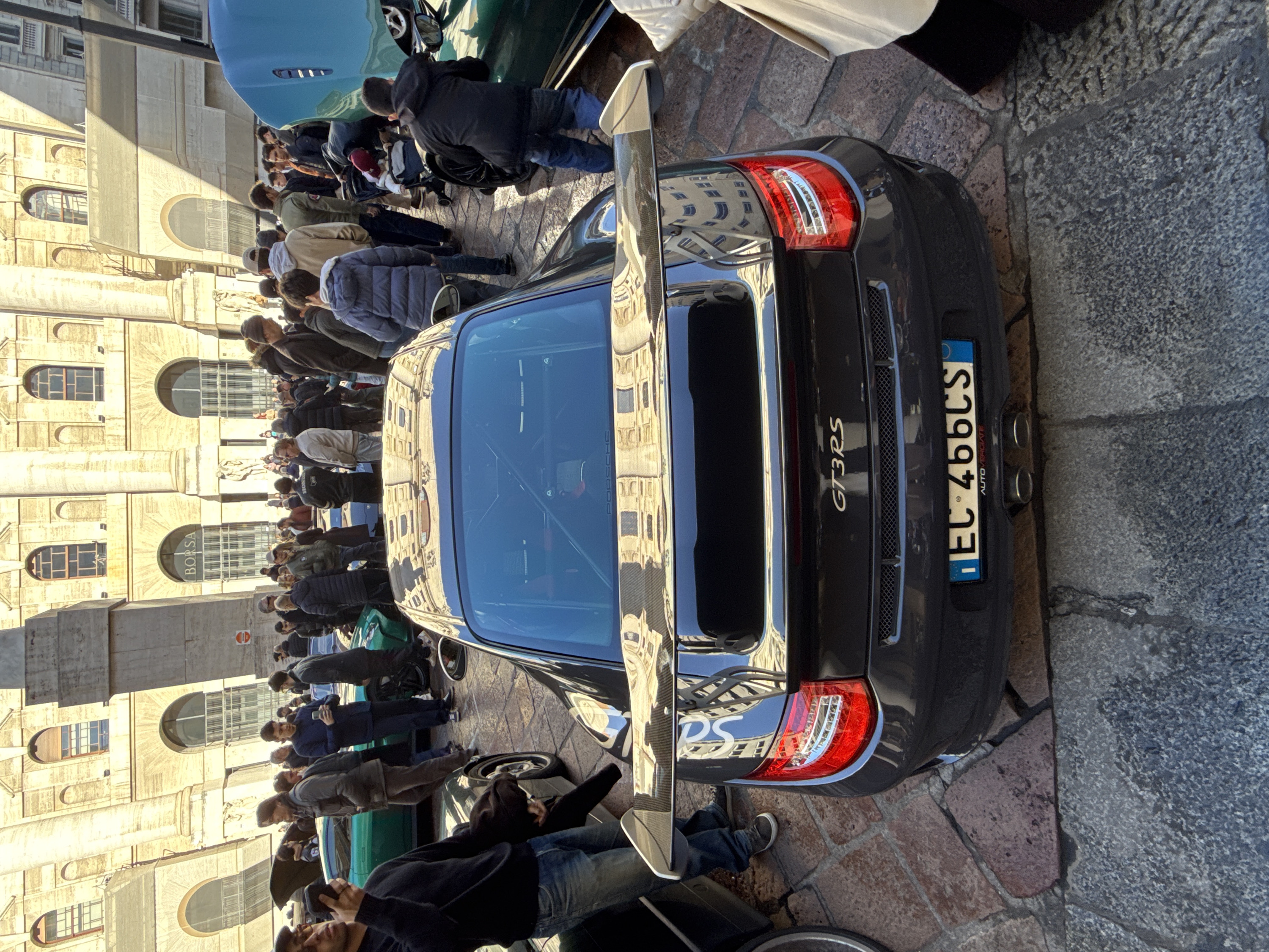 A dark grey Porsche 911 GT3 RS is centrally parked on a cobbled and paved public square, viewed from a high angle directly behind the vehicle. Its rear features a prominent large spoiler, "GT3RS" badging, red taillights, dual exhaust pipes, and an Italian license plate displaying "EC 466CS" with "AUTOVERGIATE" beneath. A multi-point roll cage is visible through the rear window.
A crowd of approximately 30 to 50 individuals, primarily adults dressed in casual winter attire, is gathered around the cars, observing them. To the immediate left of the Porsche, a portion of a green sports car is visible. To the right, a turquoise sports car with its hood raised is partially obscured by people. A young boy in a dark jacket and blue jeans kneels by the turquoise car, looking into its engine bay. Nearby, an adult pushes a baby in a stroller. On the far left, a man holds a smartphone, appearing to photograph the event.
The background is dominated by the grand, neoclassical facade of Palazzo Mezzanotte, home to Borsa Italiana, located in Piazza Affari, Milan. The building features large fluted columns, ornate arches, and rectangular windows. The word "BORSA" is distinctly visible above one of the arched ground-floor windows. Sunlight illuminates the scene, casting shadows across the building and pavement.