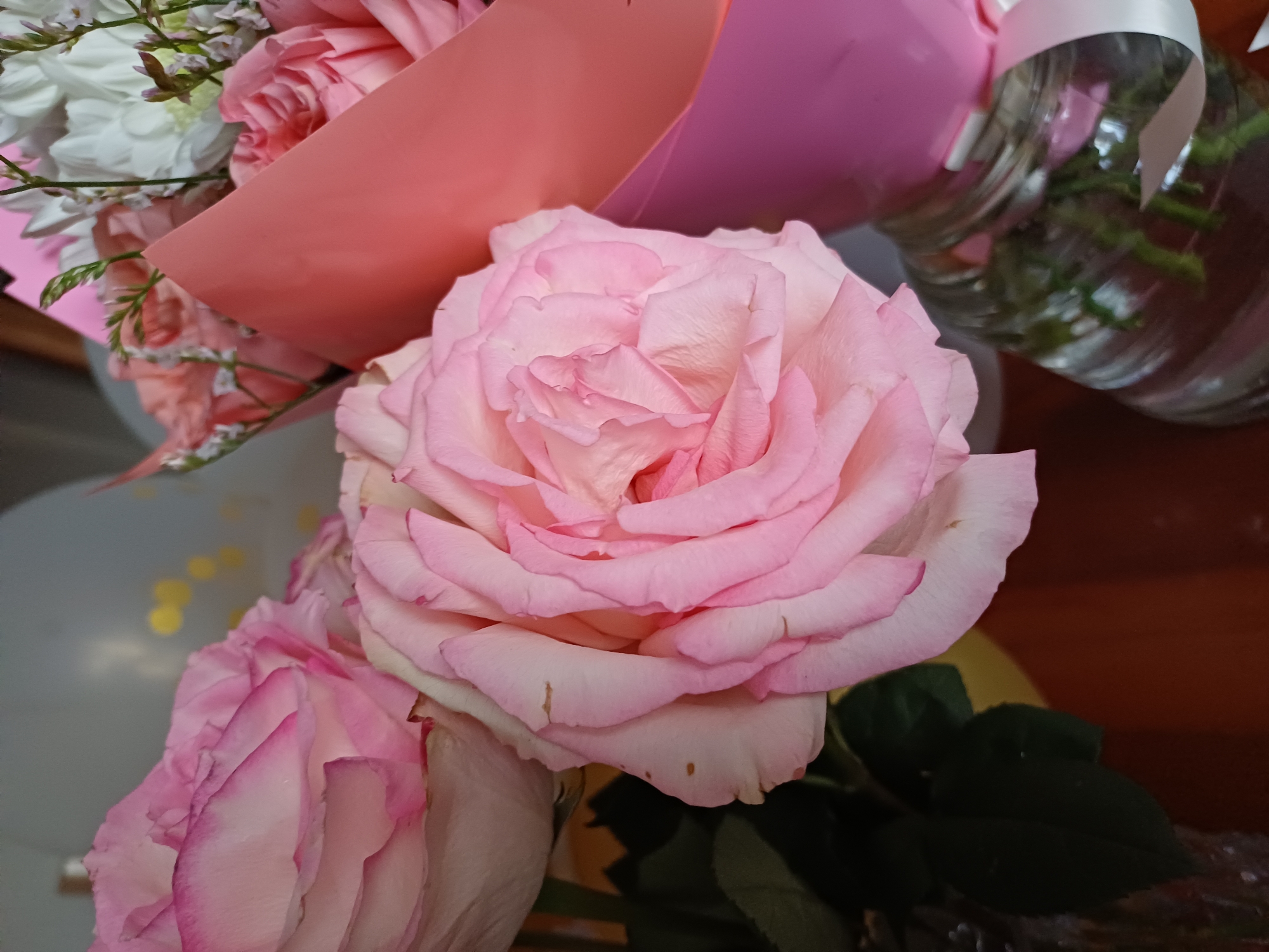 A close-up, eye-level, indoor shot focuses on a bouquet of pink roses. The foreground is dominated by a large, light pink rose with delicately ruffled petals. To the left, another pink rose, slightly out of focus, shows hints of darker pink on its edges. Behind the roses, a pink floral wrap is visible, partially obscuring other flowers and greenery in a bouquet. A clear glass vase filled with water and some stems supports part of the bouquet. The background is soft and blurred, suggesting an indoor setting with hints of light sources or decorations. The overall impression is of a gentle, floral arrangement.