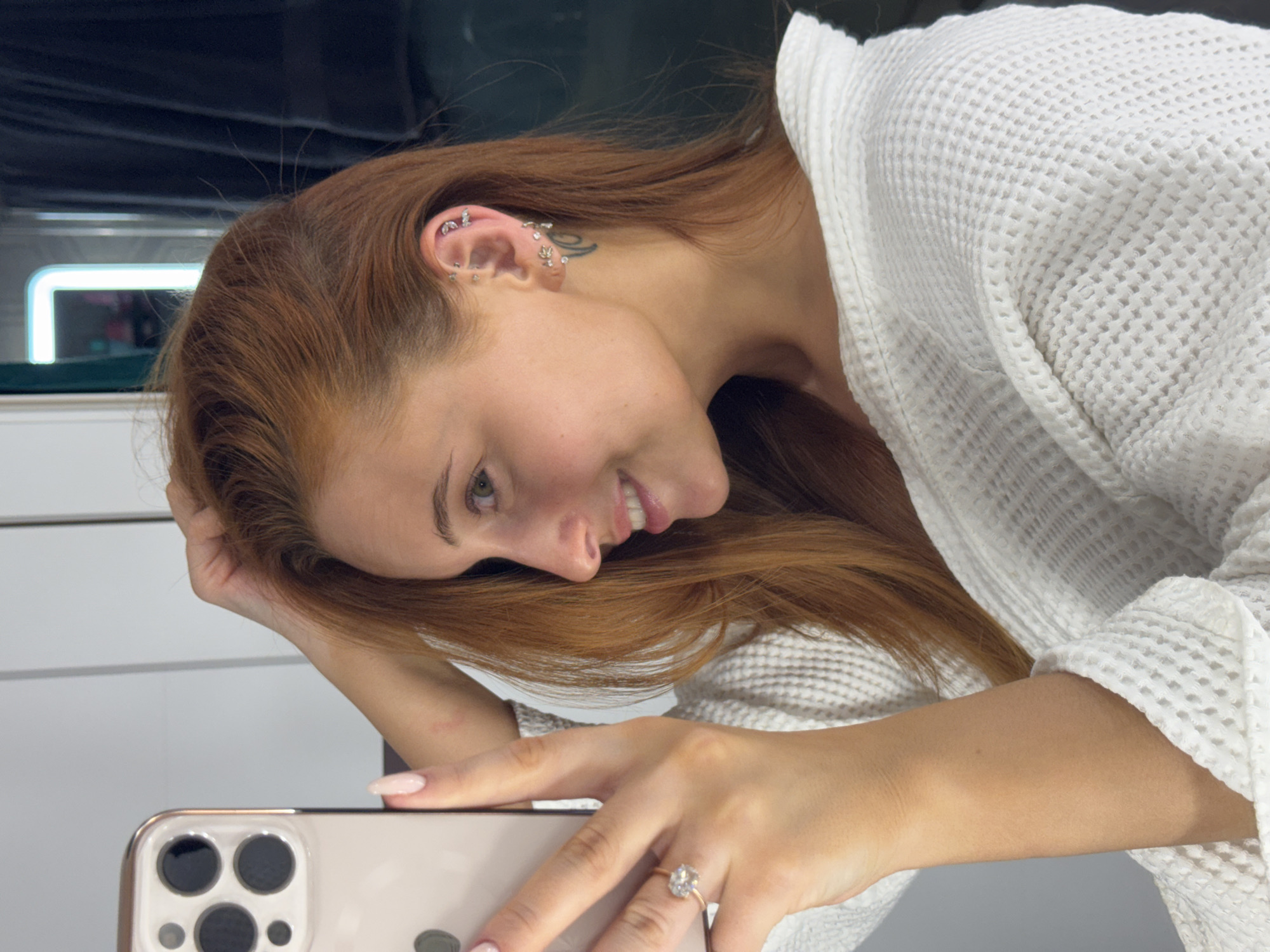 A woman with red hair, wearing a white waffle-weave robe and an engagement ring, is taking a selfie with a modern smartphone. She is indoors, likely in a bathroom, with a light-colored wall in the background. The location is Chaikovsky, Russia.