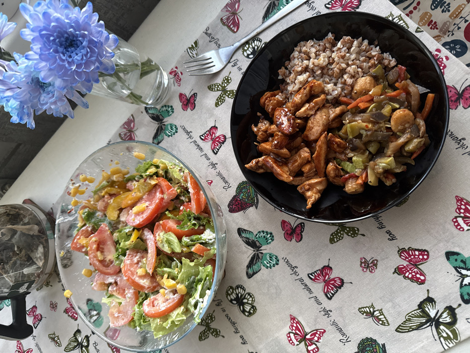 The image depicts a prepared meal laid out on a table, identified by the prompt as being in Saint Petersburg, Russia. The table is covered by a light-colored textile placemat or runner featuring a repeating pattern of multi-colored butterflies and cursive English text, including phrases such as "Romantic style and natural elegance" and "Happy day in sooting way."
In the upper left, a clear glass bowl holds a salad composed of green leaf lettuce, sliced red tomatoes, yellow corn kernels, and other diced vegetables, coated in a creamy white dressing. To the lower right, a black circular plate contains three distinct food items: glazed, brown-sauced meat pieces (consistent with chicken), a serving of cooked buckwheat groats, and a mixture of sautéed vegetables including zucchini, carrots, and onions in a light sauce. A silver metal fork rests on the placemat to the right of the black plate.
A clear glass teapot, with a black handle and a visible tea tag, sits in the upper central-left, containing brewed loose-leaf tea. In the upper right, a clear glass cylindrical vase holds several vibrant blue-purple chrysanthemum blooms in water. The immediate background beyond the table is an indistinct, light-toned surface. No individuals are visible.