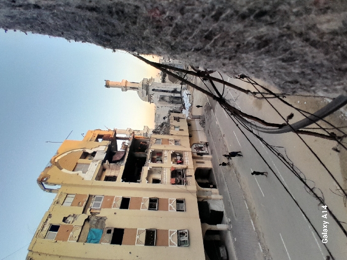 A scene shows significant destruction with a tan building bearing extensive damage, appearing bombed. The top section is severely impacted, revealing charred interiors. Adjacent stands a mosque with a tall minaret, seemingly untouched. A street runs parallel to the buildings, with several dark figures walking. Power lines crisscross overhead, adding to the chaotic atmosphere. The image is taken from a high vantage point with a blurry, gray texture at the top, possibly the edge of a structure. "Galaxy A14" is printed in the lower right corner.