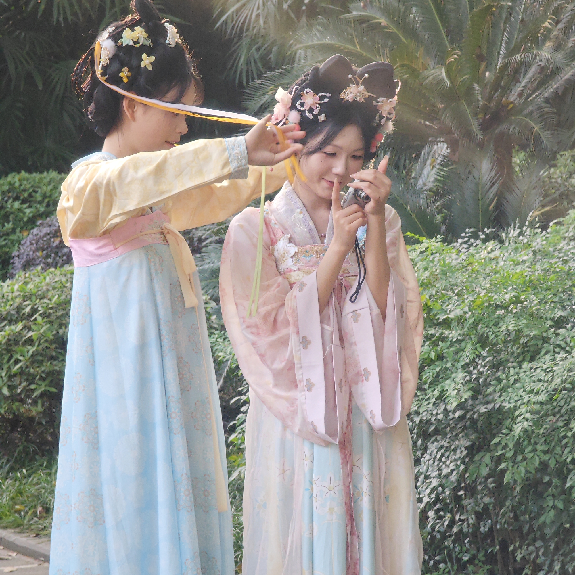 Two young Asian women are outdoors in Yuzhong District, China. One woman is adjusting a ribbon on the head of the other woman, who is holding a camera. Both women are wearing traditional Chinese clothing. The environment includes lush green bushes and a palm tree.