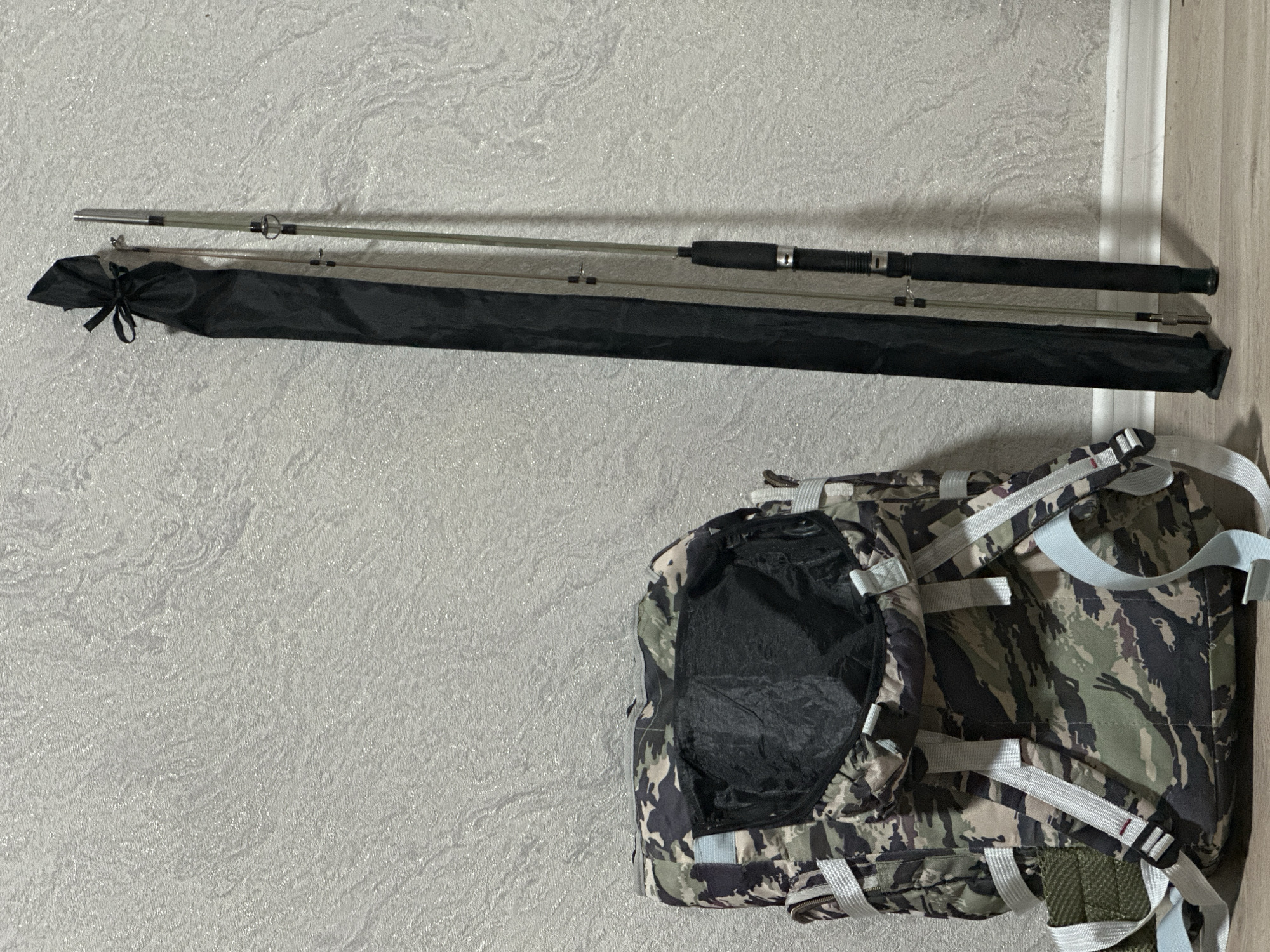 A camouflage backpack and a fishing rod in a black protective case stand upright against a textured, light grey wall with a wooden floor at the base of the wall. The backpack is positioned on the left side of the image, with its main compartment open. The fishing rod and its case are on the right side, slightly angled. The scene appears to be indoors, possibly in a storage area or a room. There is no indication of the specific location within Almaty, Kazakhstan.