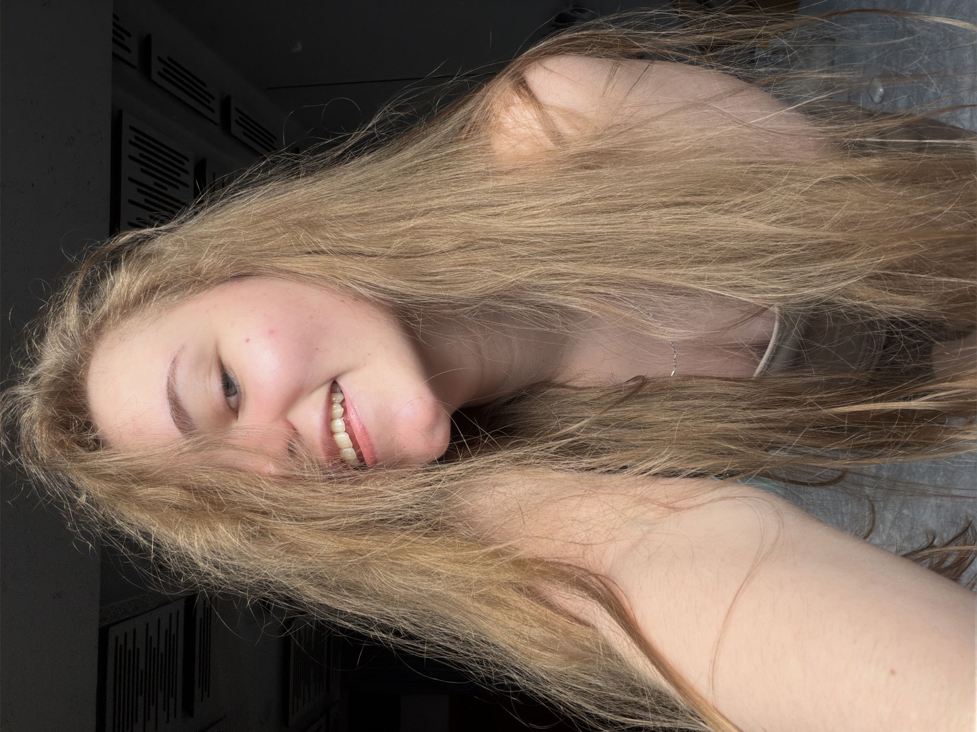 A young woman with long, blonde hair is smiling into the camera, her hair partially obscuring her face. She is wearing a light-colored tank top. The background is dark, with what appears to be a textured ceiling or wall in the upper portion of the image. The overall lighting suggests an indoor setting, possibly a residential space in Dnipro, Ukraine.