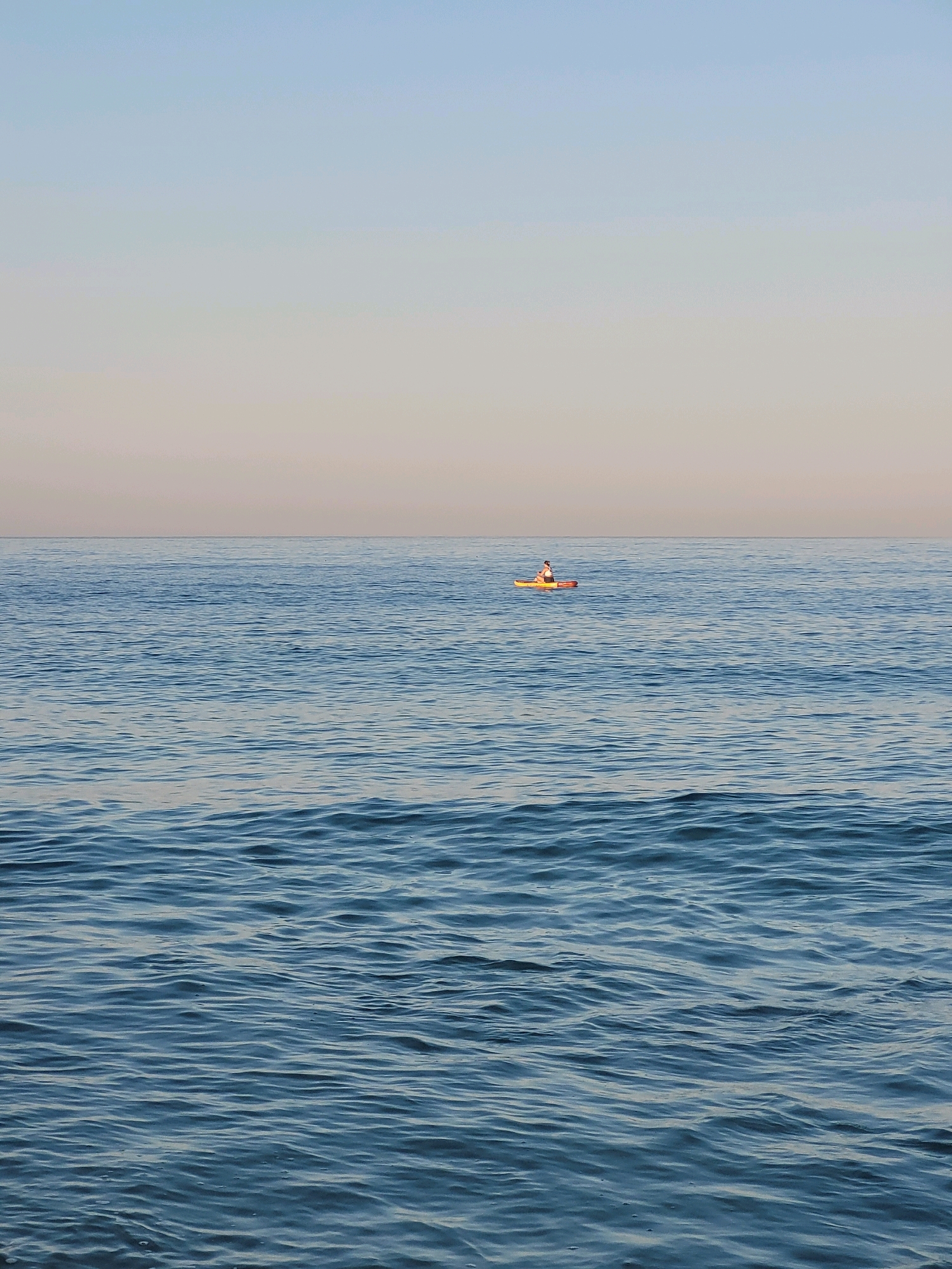 A person is paddleboarding on a calm, blue ocean. The paddleboard is yellow and orange, and the person is wearing a white shirt and a hat. The ocean extends to the horizon, where it meets a pale sky. The image is taken from a low angle, with the water filling the majority of the frame. The location is not specified in Alanya, Turkey, as the image does not provide any visual cues to determine the location.