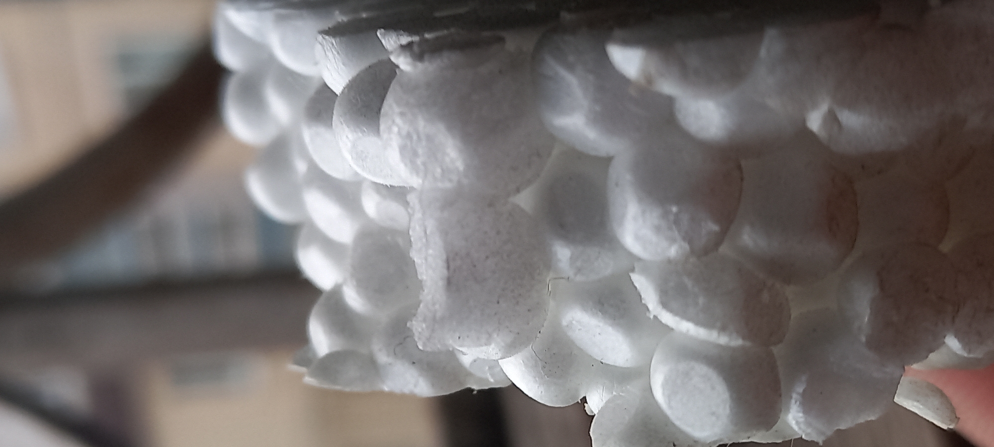 A macro photograph depicts a segment of white expanded polystyrene (styrofoam) material, observed in Kryvyi Rih, Ukraine. The material is composed of numerous fused, predominantly spherical beads, presenting a porous and irregular surface texture. Visible on the material are indications of breakage and tearing, particularly along the edges. Scattered dark particulate matter and localized light brown discoloration are present on various polystyrene beads. The background is extensively blurred, showing indistinct, out-of-focus shapes consistent with urban or architectural elements, illuminated by natural daylight. No individuals, specific actions, or interactions are discernible within the frame.