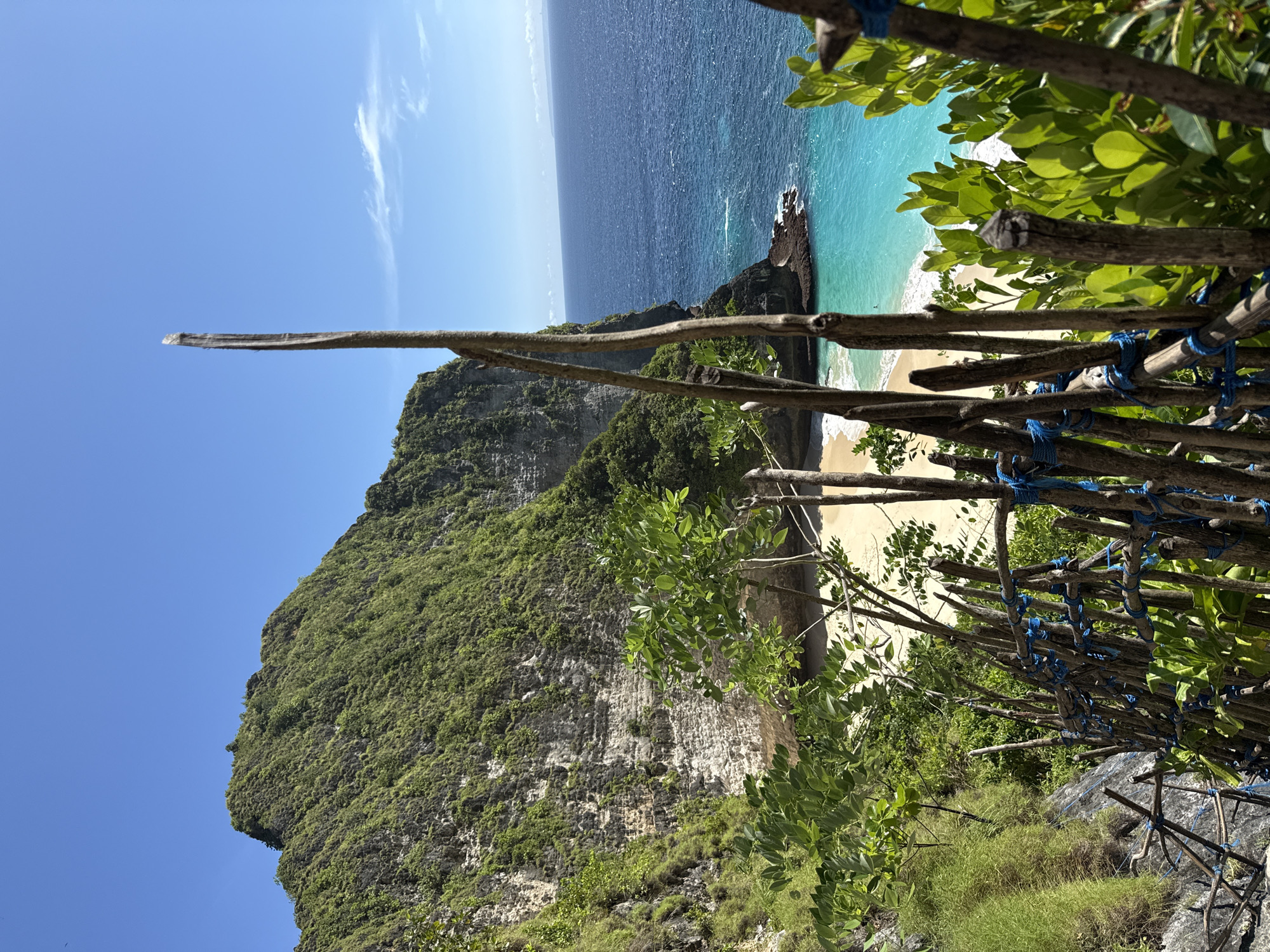 A landscape photograph taken in Bunga Mekar, Indonesia, shows a cliff overlooking a beach and ocean. The cliff face is covered with greenery. A stick fence is in the foreground on the right side. The ocean is blue and turquoise near the beach. The sky is blue with a few clouds. A long stick is pointing upward from the left.