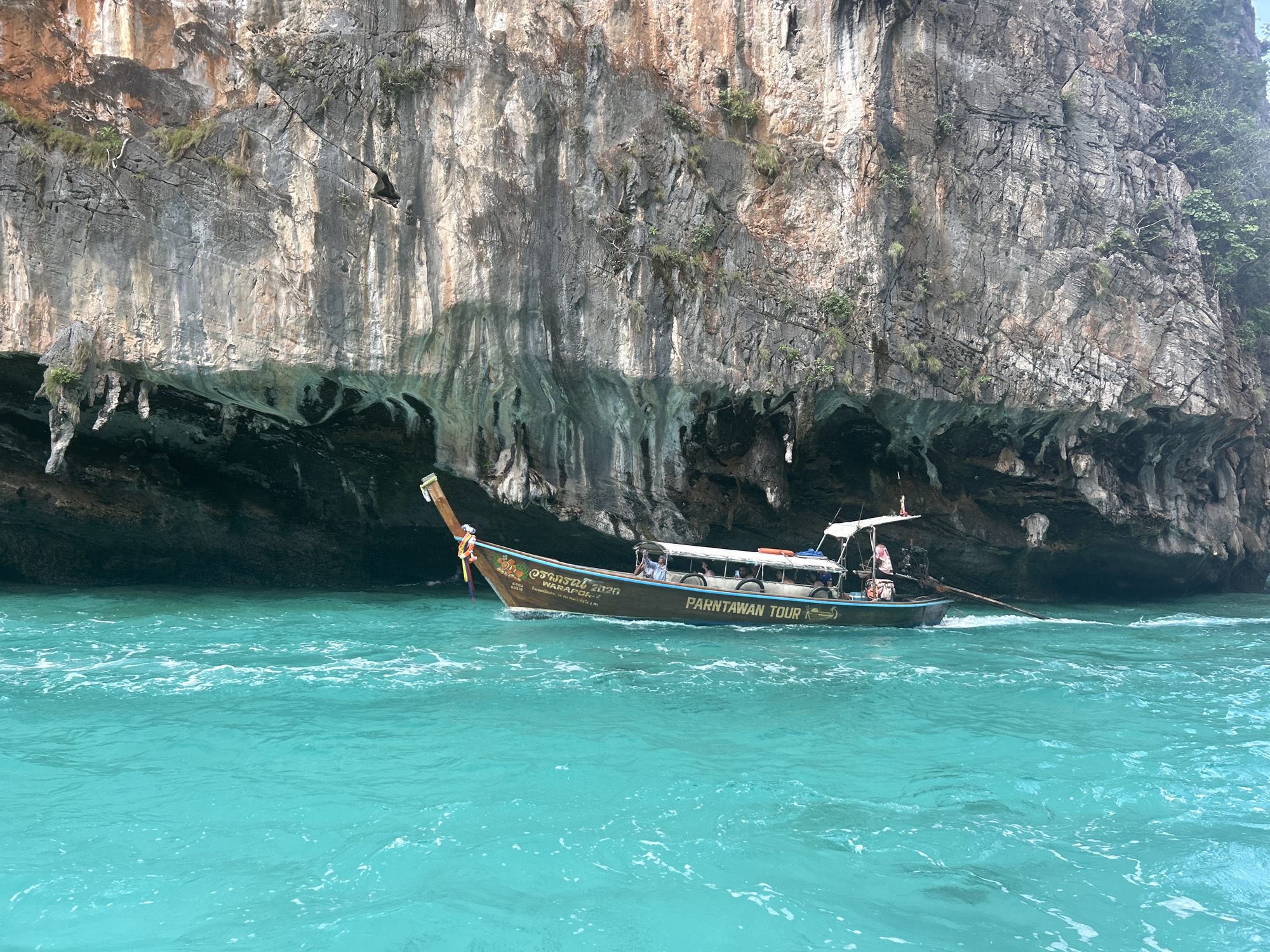 A longtail boat, identified by the text "PARNTAWAN TOUR" on its side, navigates through turquoise water next to a towering limestone cliff face. The boat, carrying multiple passengers and a captain, is positioned towards the right of the frame, with its prow pointing left. The cliff, characterized by its rough texture, varying shades of gray and brown, and patches of greenery, forms a dramatic backdrop. The water exhibits a vibrant, almost luminous turquoise hue, with white foam indicating the boat's movement. Sunlight illuminates the scene, casting shadows and highlighting the textures of both the rock and the water. The location is Haad Yao, Thailand, a region known for its stunning coastal scenery and limestone karst formations.
