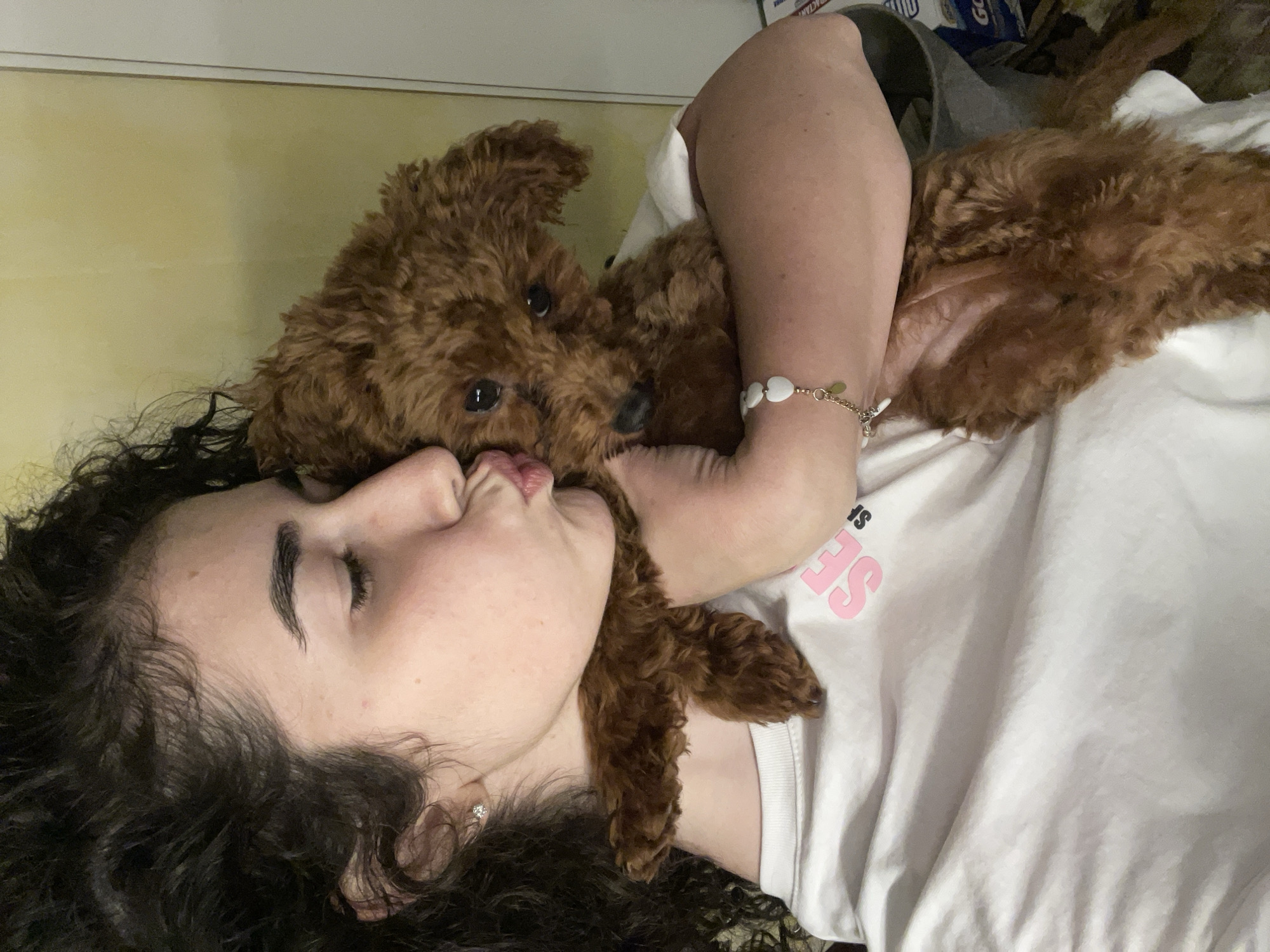 A woman with dark curly hair is lying down, with a brown poodle lying on top of her. The woman's eyes are closed, and she is puckering her lips as if to kiss the dog. She is wearing a white t-shirt, and a bracelet with white beads is visible on her arm. The dog is lying on its back with its head resting on the woman's shoulder. The background is a yellow wall and some blurred objects. No information suggests the photo was taken in Sofia, Bulgaria.