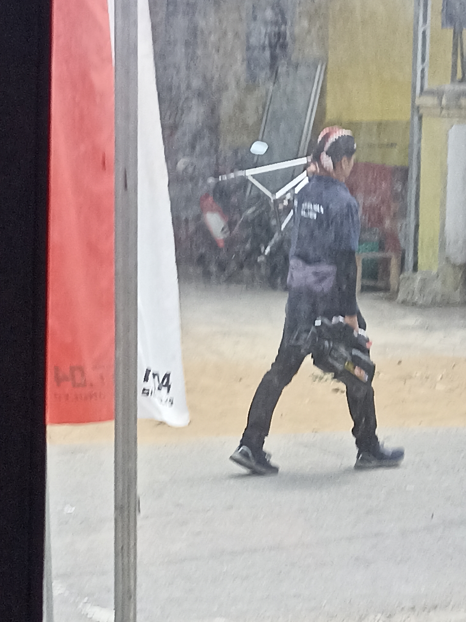 A person walks with a video camera in Pulisen, Indonesia. The person, who is wearing a dark blue shirt and black pants, is carrying a black video camera in their right hand. They are walking from left to right across a sandy surface. In the background, there is a red and white banner, a yellow building, and a motorcycle. The image is taken from a slightly elevated angle.