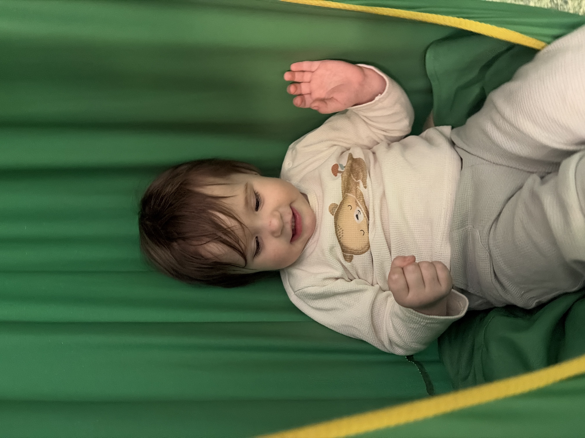A baby lies in a green hammock, likely indoors, as the location is not discernible from the image. The baby is wearing a long-sleeved white shirt with a brown giraffe design and light gray pants. The hammock has yellow trim. The baby is lying on their back, smiling, with one hand open and the other in a fist.
