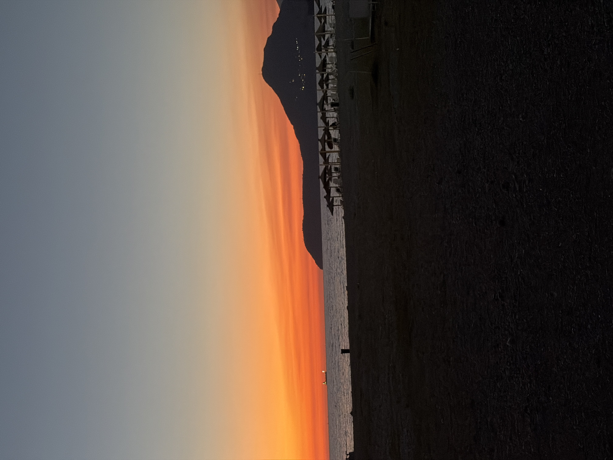 This is a vertical, wide shot of a beach at sunset. The sun is setting behind a dark, silhouetted mountain range that stretches across the frame. The sky is a gradient of soft grays on the left, transitioning to vibrant oranges and yellows towards the right, creating a dramatic sunset effect. The ocean is visible as a calm, dark body of water reflecting the colors of the sky.
On the dark beach, there are rows of unoccupied beach chairs and umbrellas, suggesting a resort or public beach area that is winding down for the day. The sand appears dark, possibly due to its composition or the lighting conditions. In the distance on the water, a few small lights are visible, which could be boats or structures.
The overall mood is serene and peaceful, capturing the transition from day to night in a coastal setting. The time of day is clearly sunset, with the warm colors of the sky being the dominant feature. There are no visible people or text in the image. The location context provided indicates this scene is in Bar, Montenegro.
