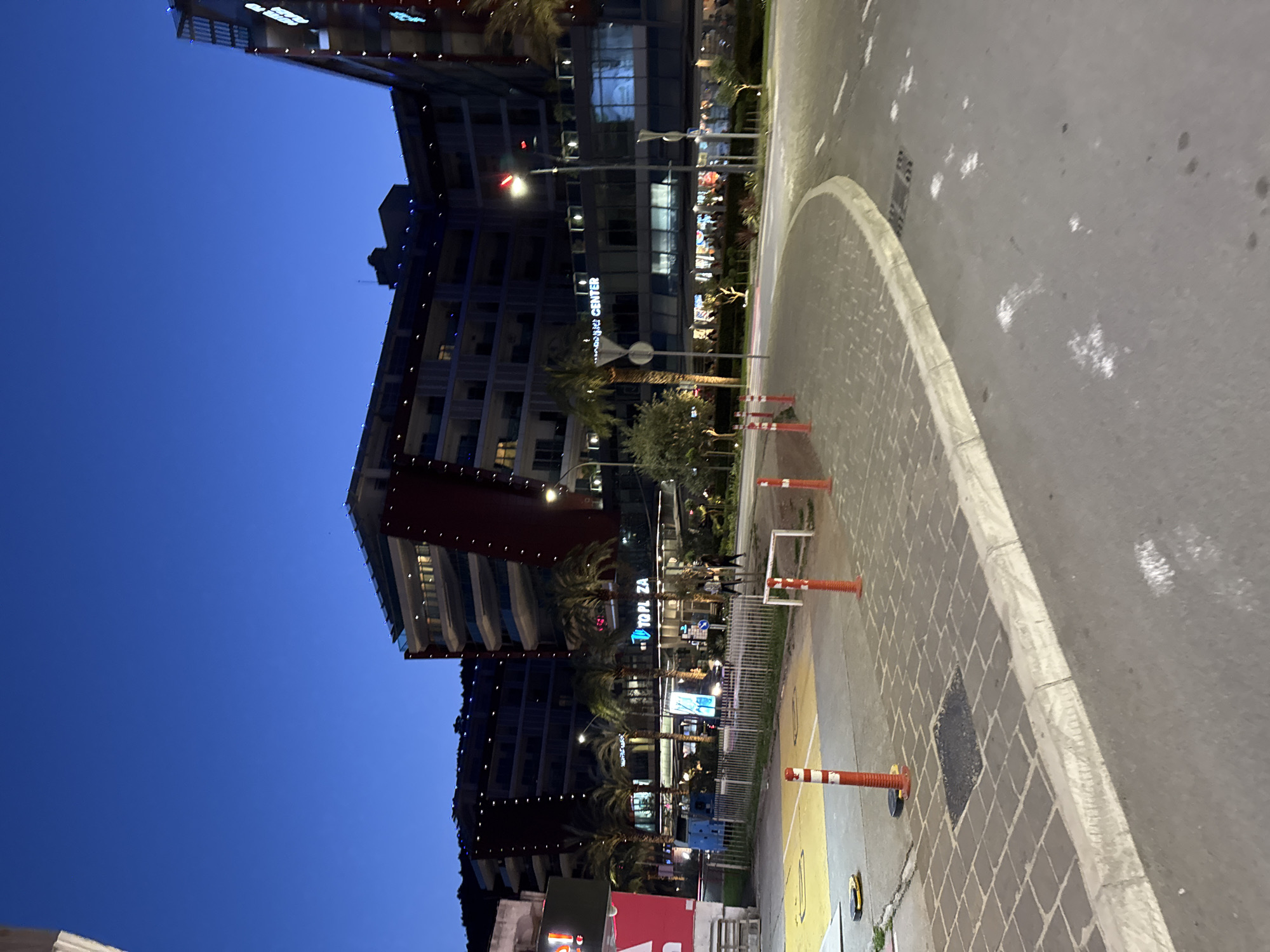 The image depicts a nighttime scene in Budva, Montenegro, showcasing modern architecture. Buildings are illuminated with lights, and palm trees line the street. Visible signage includes "Topla Za" and "Budva Center". Orange and white traffic cones are placed along a paved area. The sky is a deep blue.