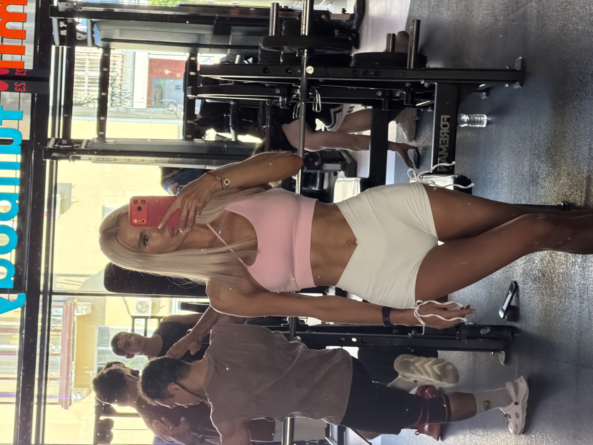 The image depicts a woman taking a mirror selfie in a gym. She is wearing a pink sports bra and white shorts, with blonde hair and visible abs. In the background, other gym-goers are visible, some engaged in workouts. The gym appears to be modern, with weightlifting equipment. The environment suggests a public space. The presence of a gym in Kazan, Russia, would indicate it is part of the city's fitness infrastructure.