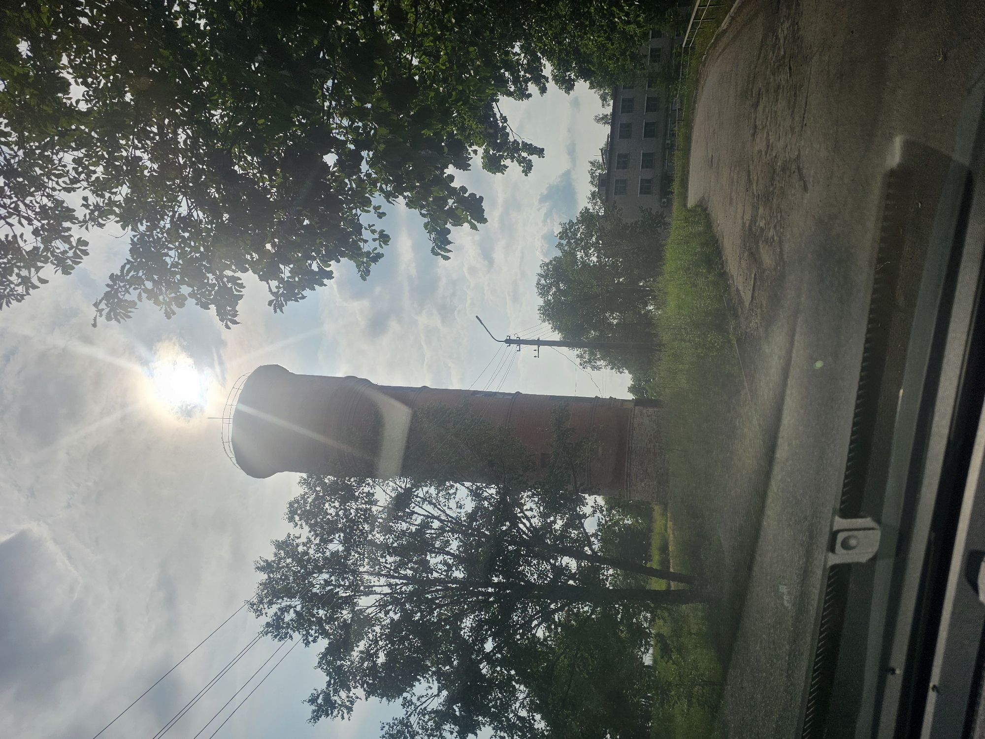 The image captures a view from a vehicle, looking out at a brick water tower against a bright, cloudy sky with the sun directly overhead, creating lens flare. The tower, an old industrial structure, is partially overgrown with vegetation. To the left of the tower is a large tree, and to the right, a section of a multi-story building with numerous windows is visible, along with more trees. A paved road curves through the scene, leading towards the building. Power lines stretch across the sky above the tower and a lamppost stands to the left of the road. The foreground shows the interior of a vehicle's dashboard and windshield. The environment appears to be rural or on the outskirts of a town, possibly in Таёжное, Russia, given the architectural style of the building and the presence of the water tower. There are no people or animals visible.
